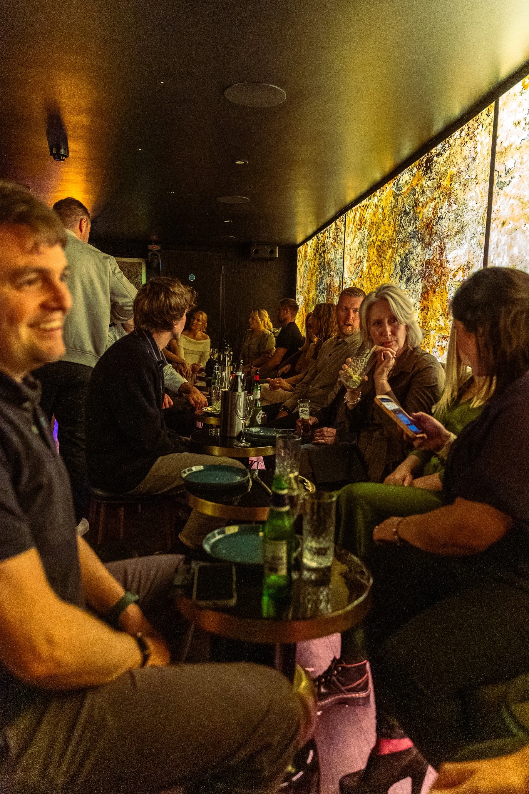 People sitting and socializing in a dimly lit bar or restaurant with a large colorful abstract mural on the wall, some individuals holding drinks, and bottles of beverages on small round tables.