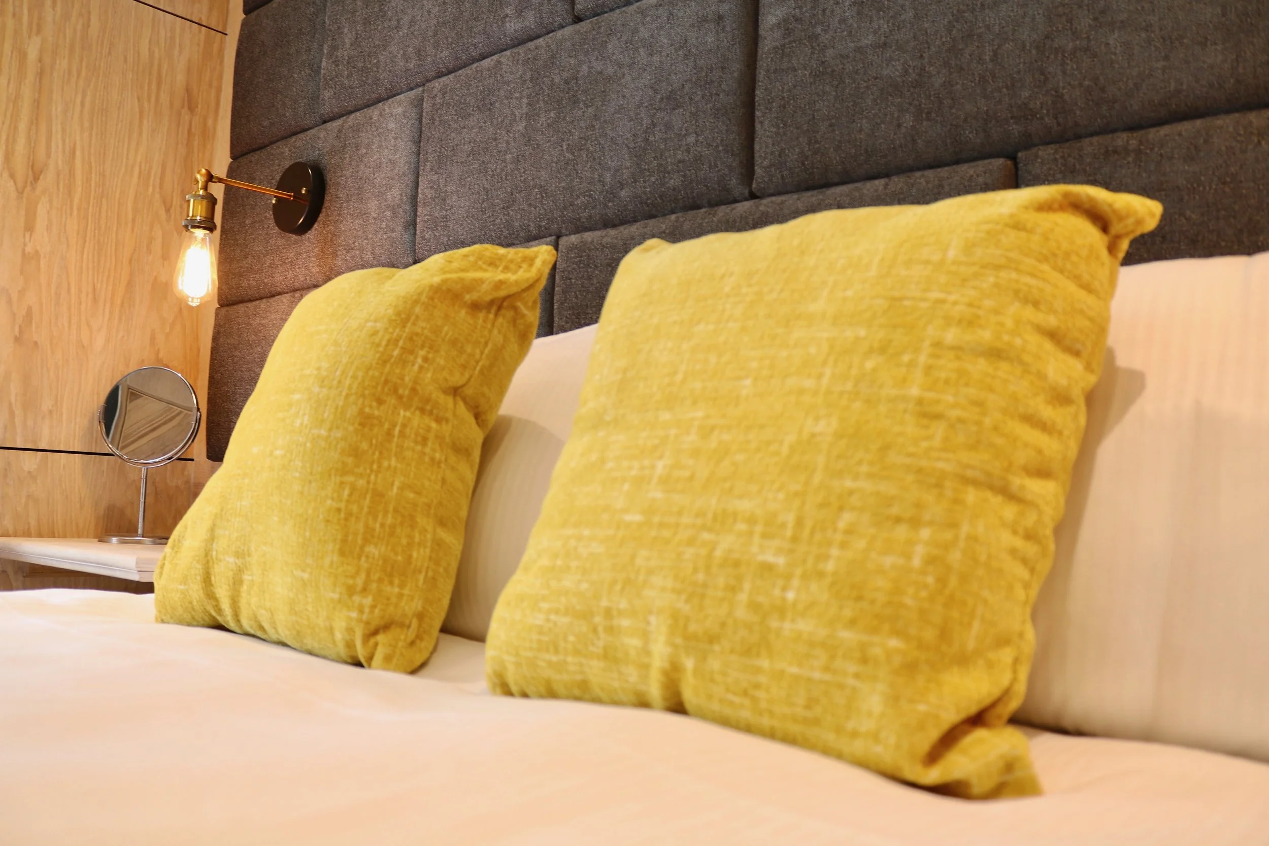 A bedroom with yellow pillows on a bed, a side table with a mirror, and an industrial-style wall lamp on a wooden wall.