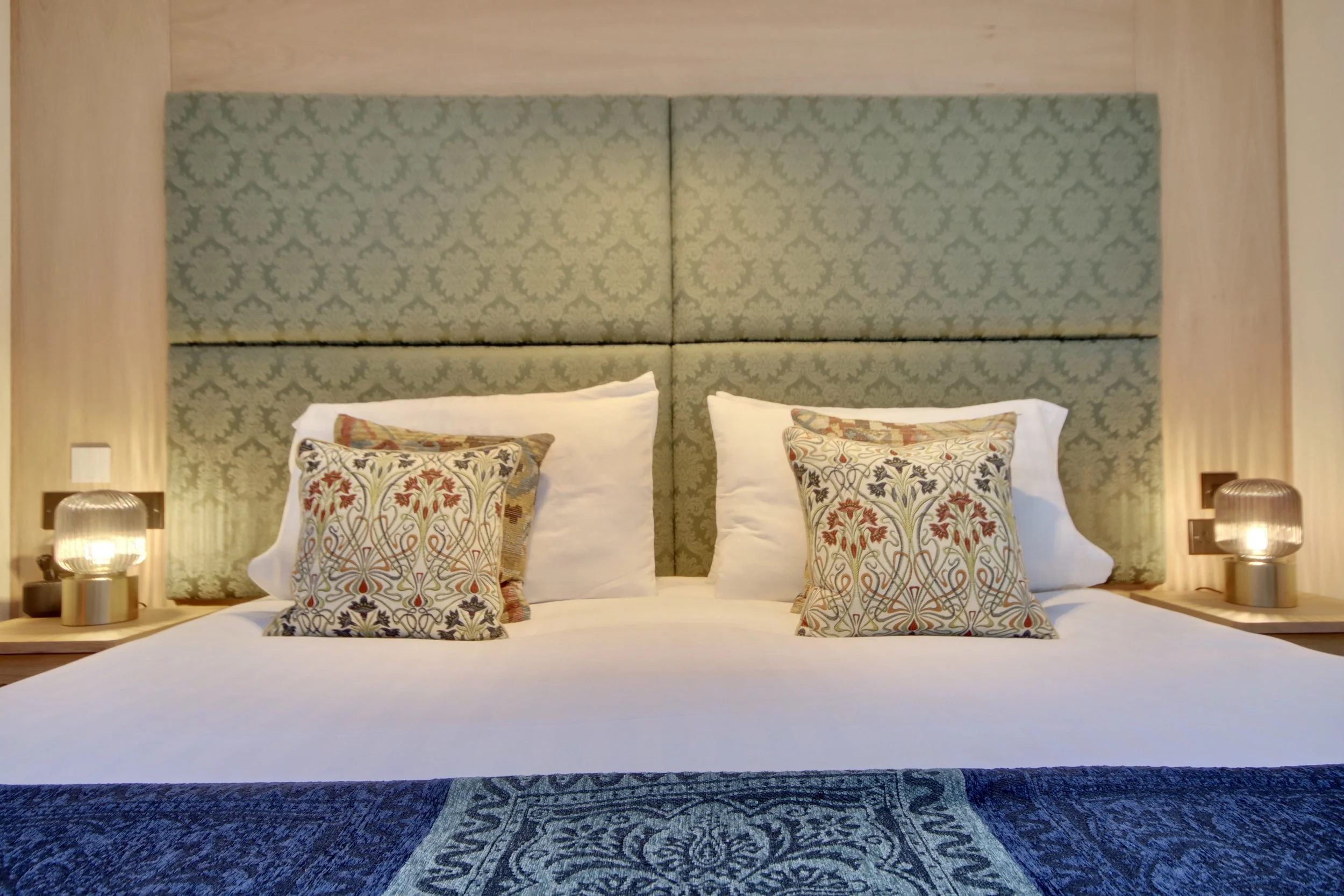 A neatly made hotel bed with white linens, four decorative pillows with floral and geometric patterns, a padded headboard with a green damask pattern, and two small round lamps on wooden nightstands on either side.