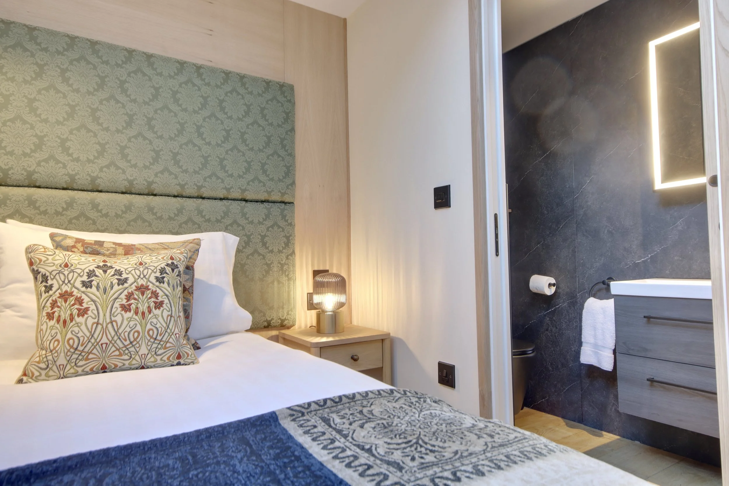 A bedroom with a bed that has patterned pillows, a wooden nightstand with a modern lamp, and a view into a bathroom with black marble walls, a mirror with LED lighting, and a towel on the floor.