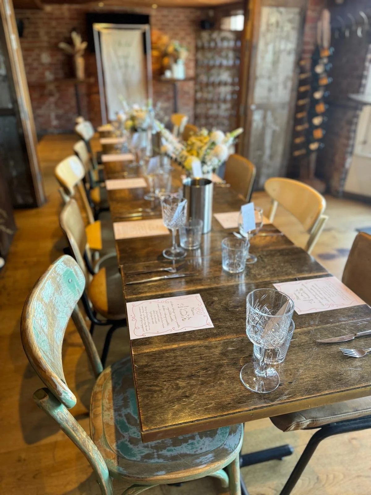 A long wooden dining table set with glasses, silverware, and printed menus, with floral centerpieces in vases. The table is in a rustic, cozy room with brick walls and wooden shelves.