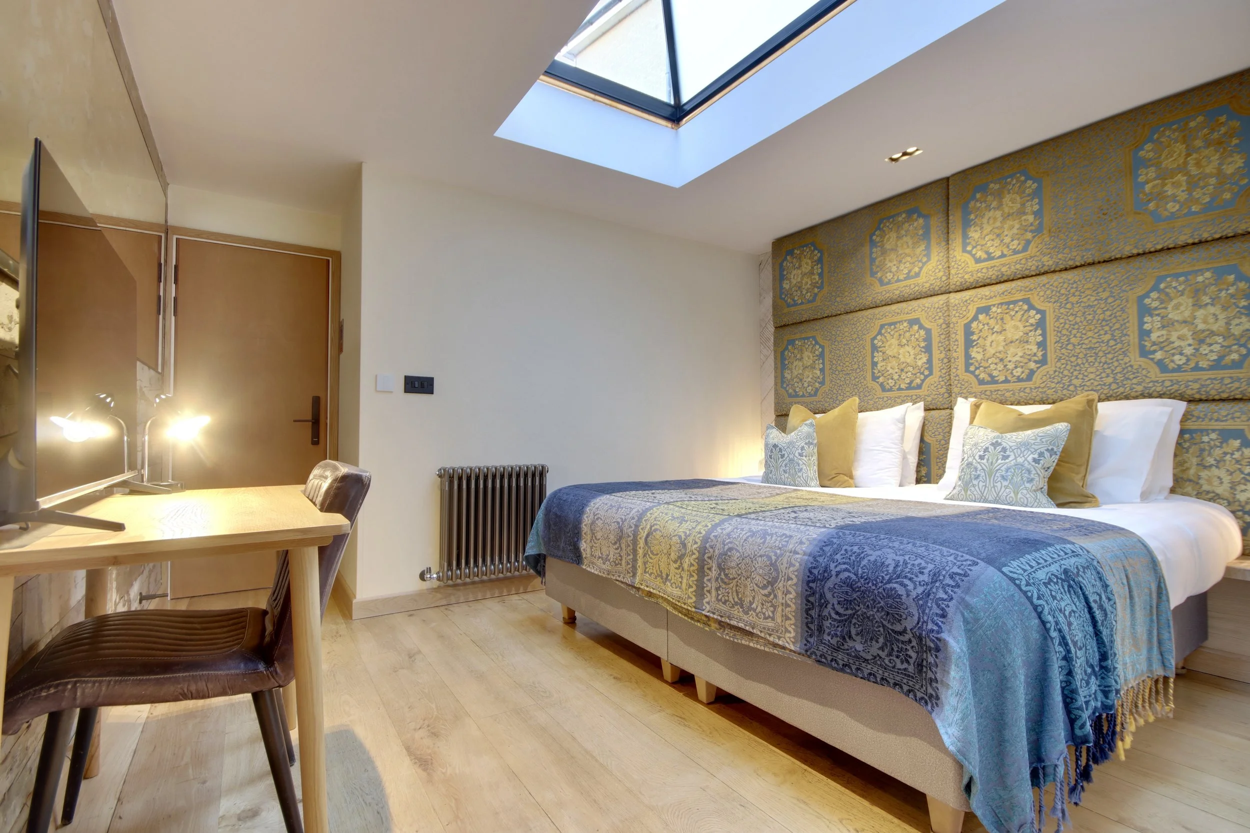 A bedroom with a bed featuring a blue and gold patterned headboard, yellow and white pillows, and a blue bedspread. A skylight in the ceiling allows natural light into the room. To the left, there is a wooden desk with a modern lamp and a dark brown chair, along with a wall-mounted radiator below. The door and light-colored wooden flooring are also visible.