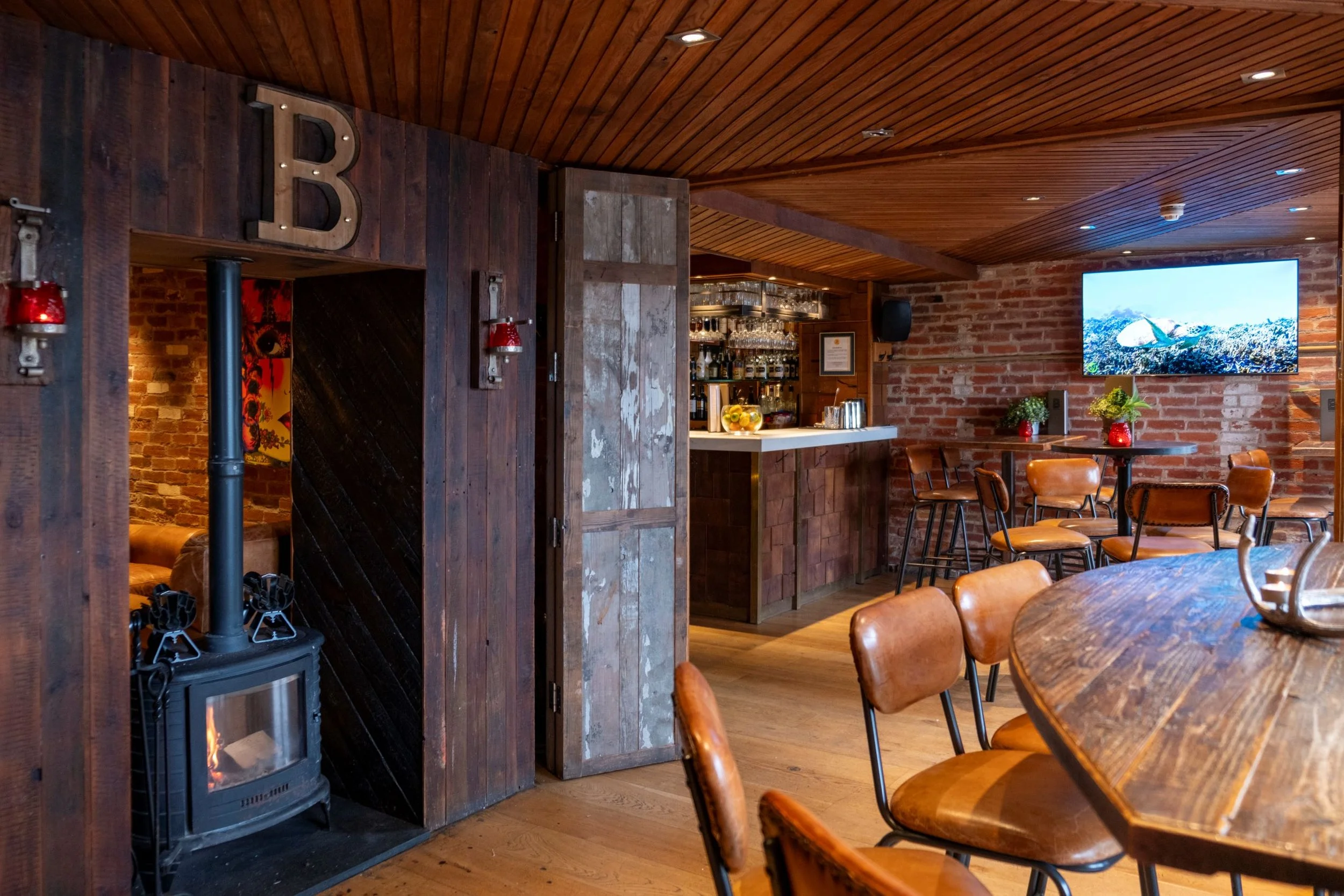 Interior of a cozy restaurant or cafe with wooden ceiling and brick walls. There is a large flat-screen TV on the wall, a bar area with glasses and bottles, and tables with leather chairs decorated with plants.