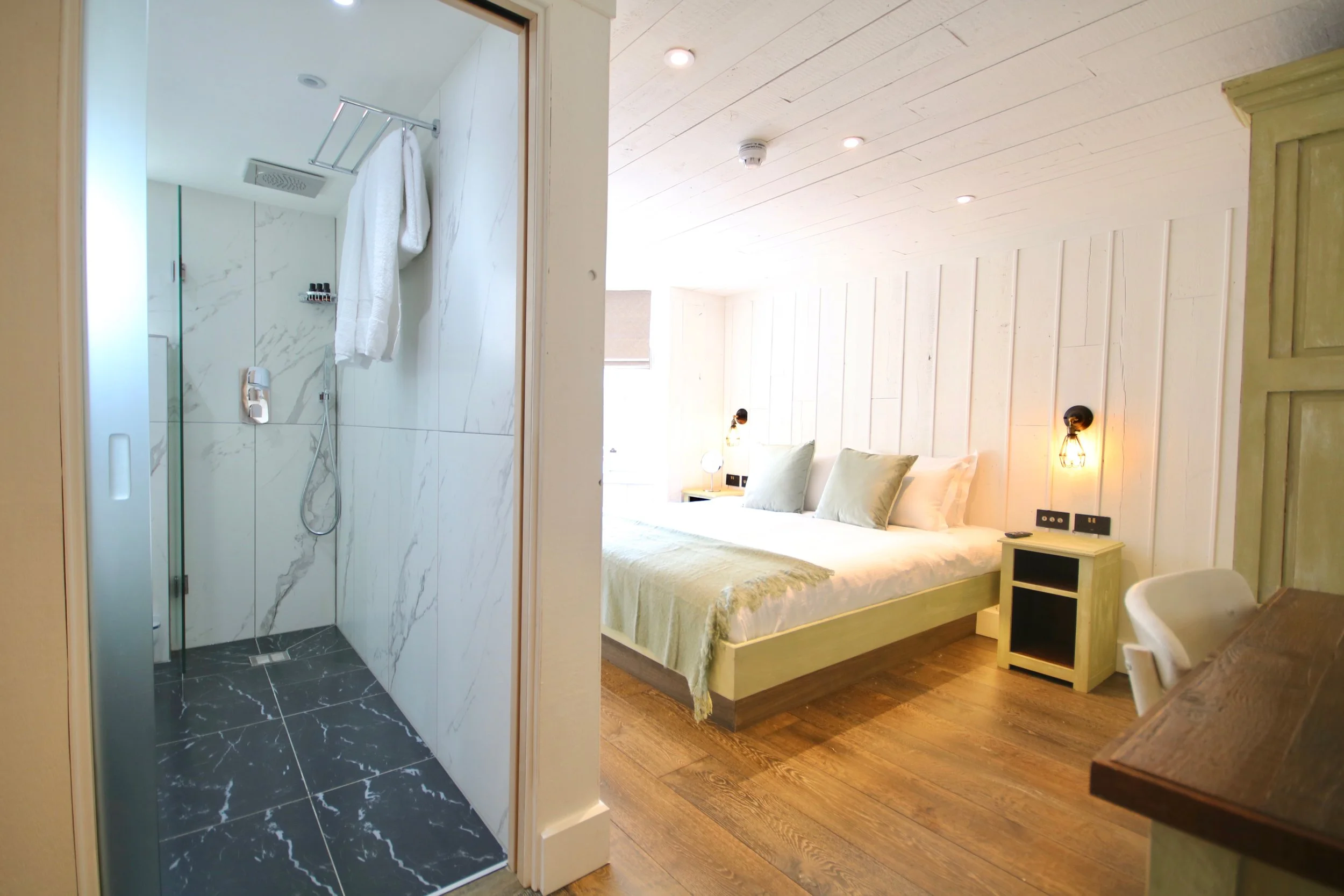 A view of a hotel room with a bed, bedside table, and lighting. To the left, there's a doorway leading to a bathroom with a glass-enclosed shower, white marble walls, and black marble flooring.