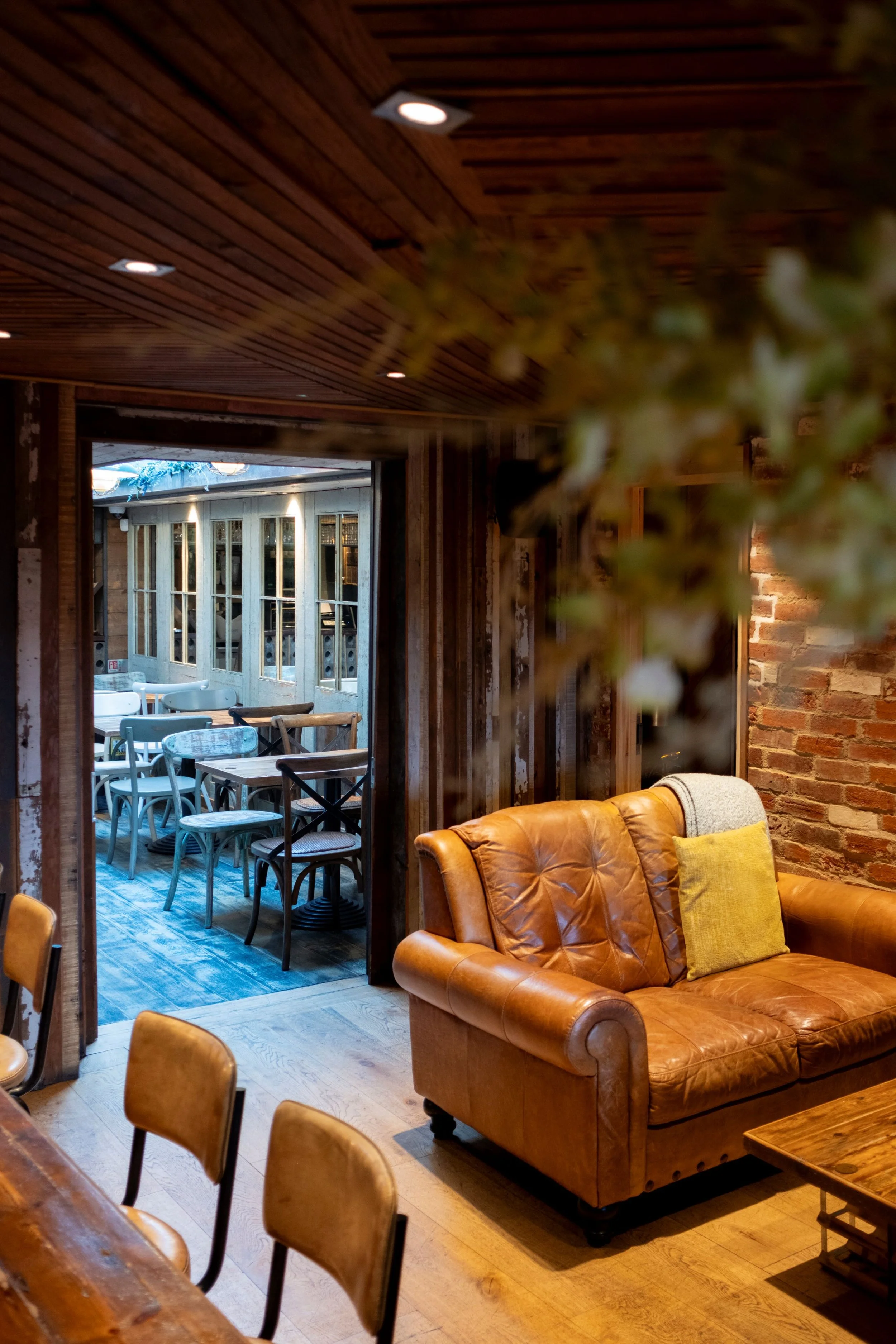 A cozy indoor seating area featuring a brown leather sofa with a yellow pillow, wooden chairs, and a view through a doorway to an outdoor patio with additional chairs and tables. The interior has warm wooden walls and ceiling with recessed lighting, 