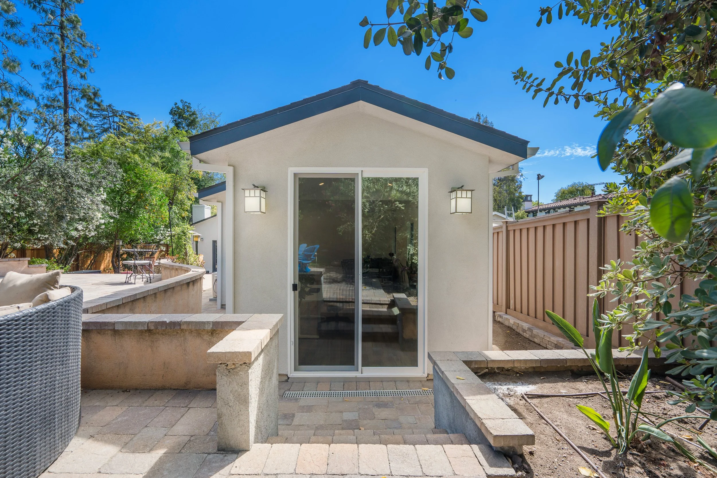 Backyard with patio, built-in seating, sliding door leading inside, two outdoor wall lanterns, wooden fence, trees, and blue sky.