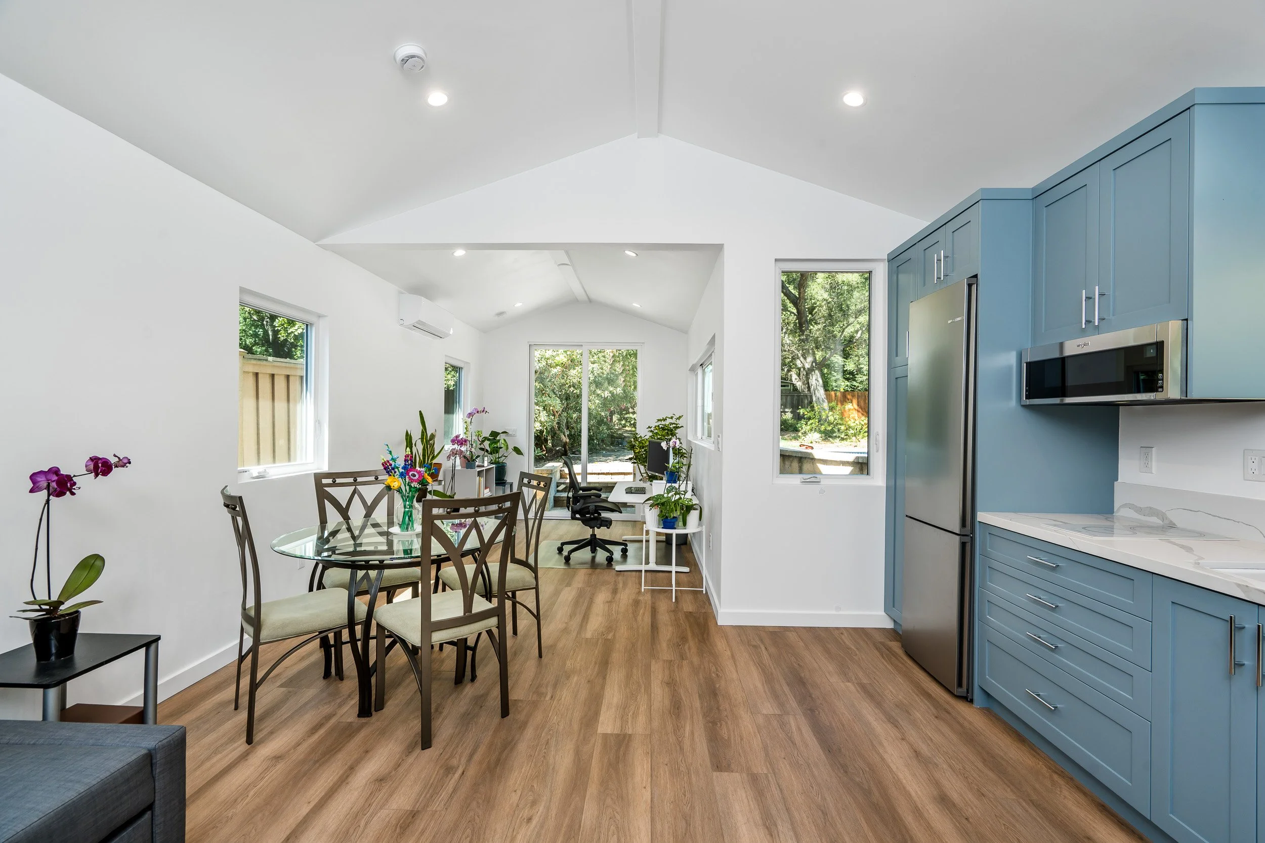 Modern kitchen and dining area with white walls, light wood flooring, and blue cabinetry. A dining table with six chairs, decorated with colorful flowers. Multiple large windows with green trees outside, and a workspace with a computer chair near the