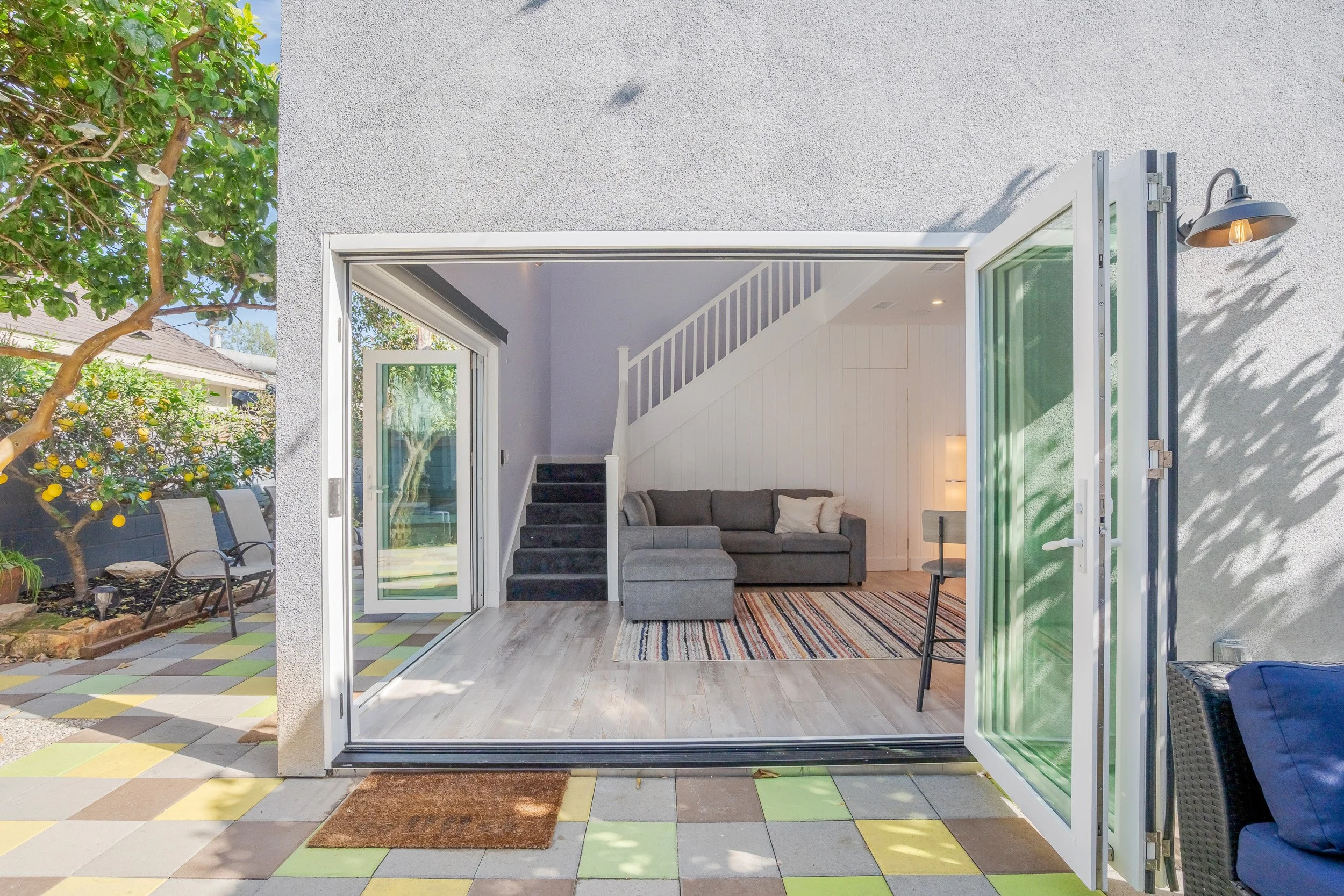 A view of a cozy outdoor patio area with a large open glass door leading into a living room. The patio has colorful checkered flooring, garden furniture, and a lemon tree outside.