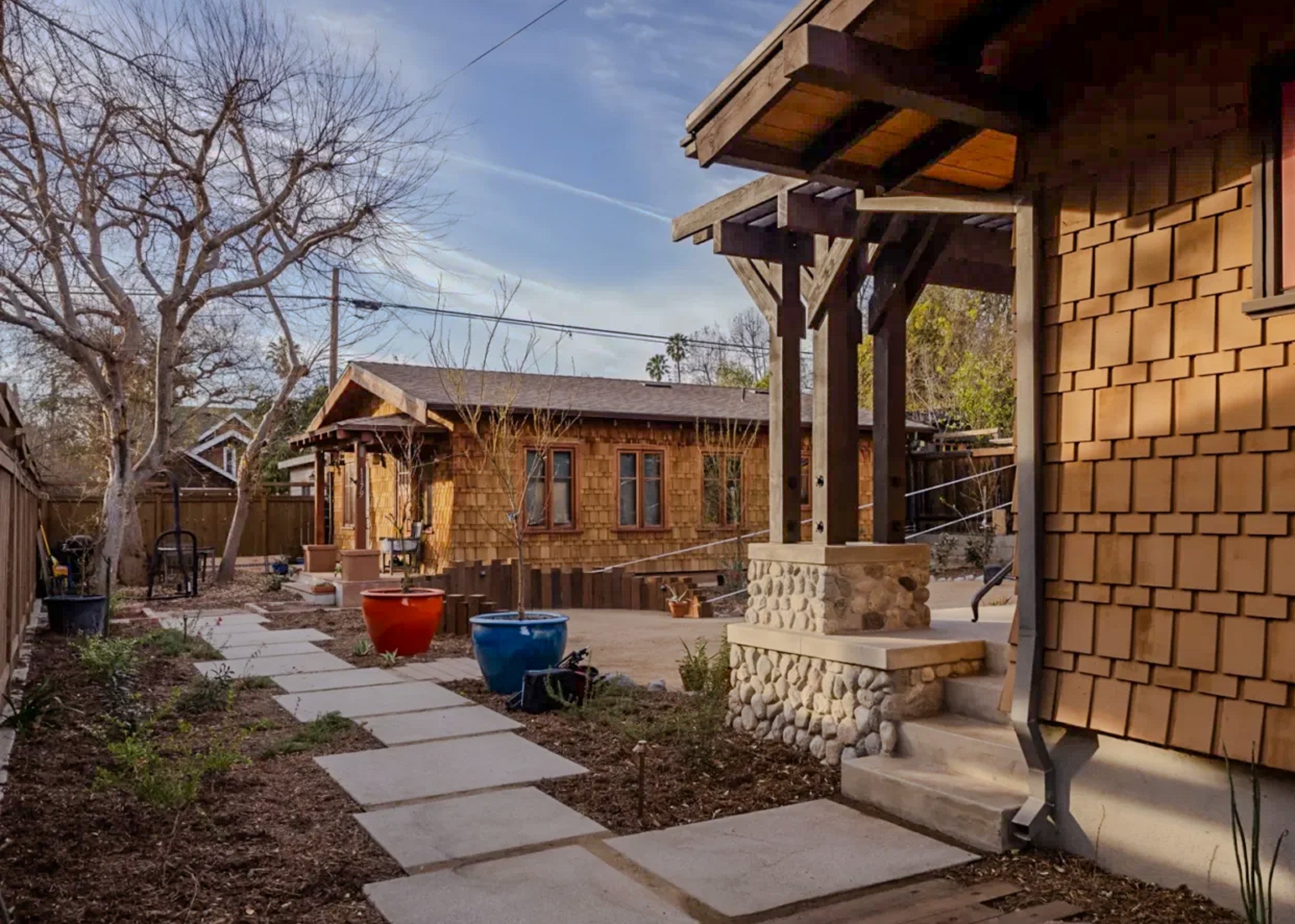 A backyard patio with a stone pathway, trees, and a wooden house with shingle siding and a front porch with stone base. There are colorful large pots and outdoor furniture, with a partly cloudy sky overhead.