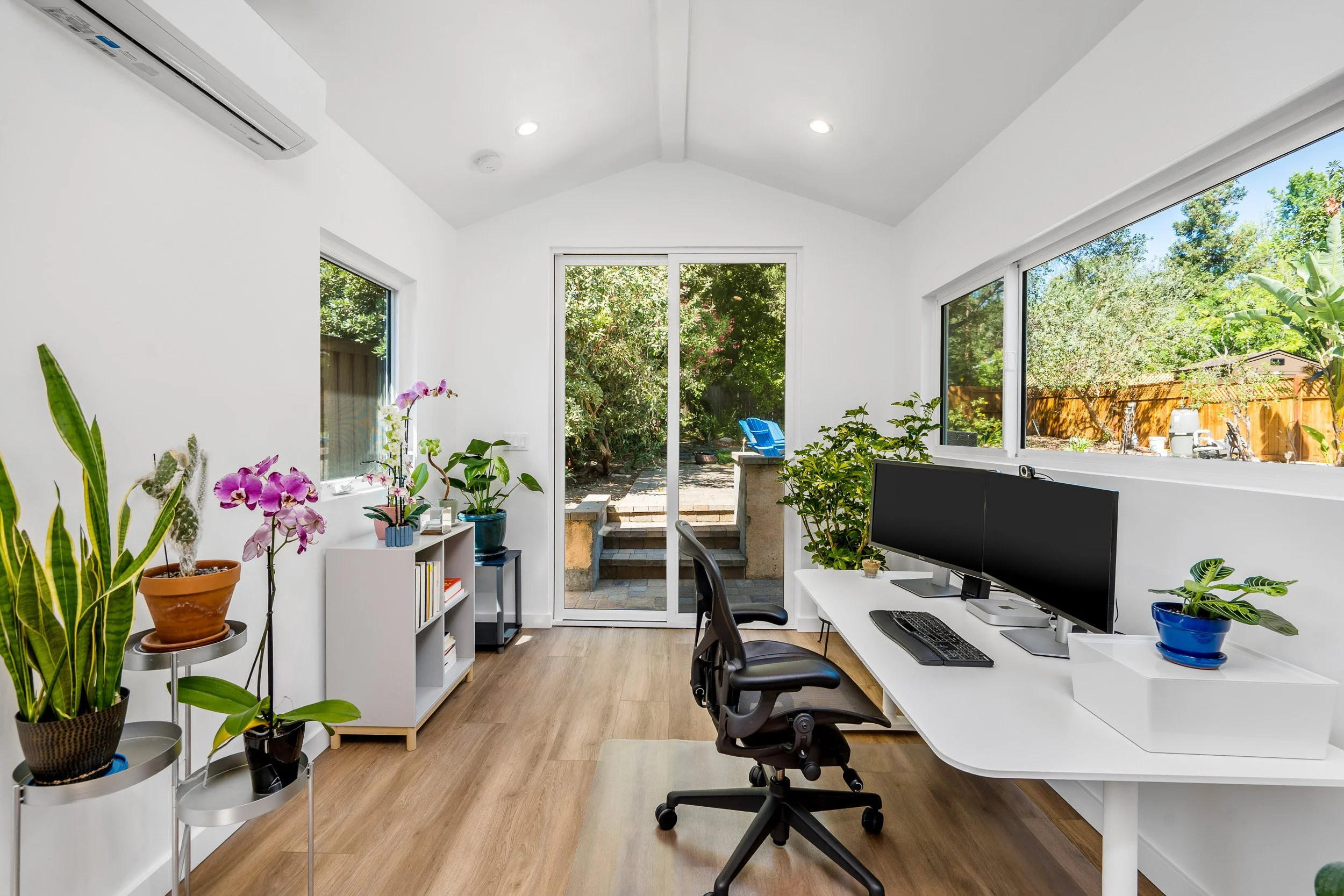 Home office with white walls, several indoor plants, a window showing an outside garden, a desk with a computer, and a sliding glass door leading to a backyard with outdoor seating.