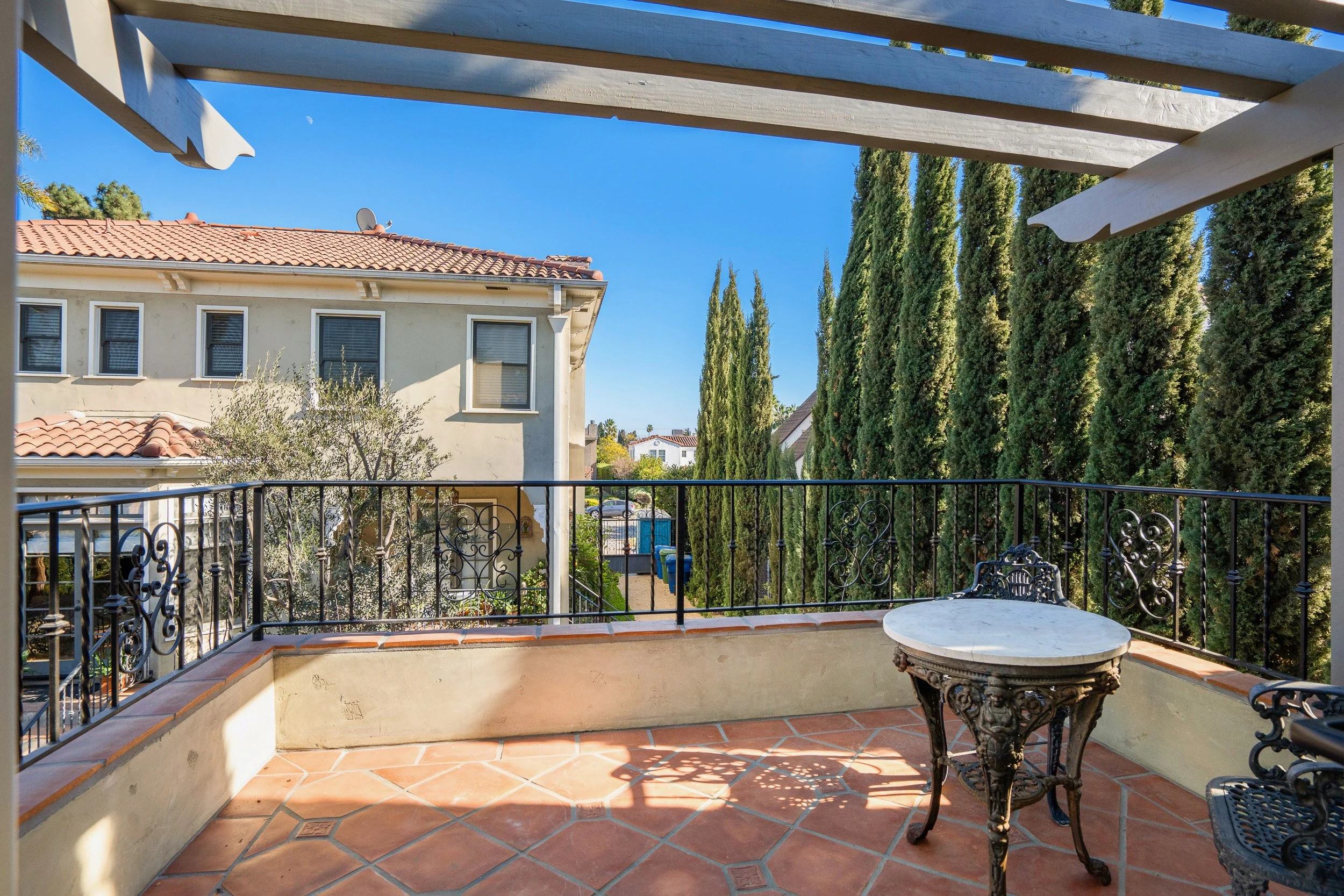 View of a balcony or patio with terracotta tile flooring, ornate black metal railing, and a small round marble-topped table. The background includes a beige residential building with a tile roof, tall cypress trees, and a clear blue sky.