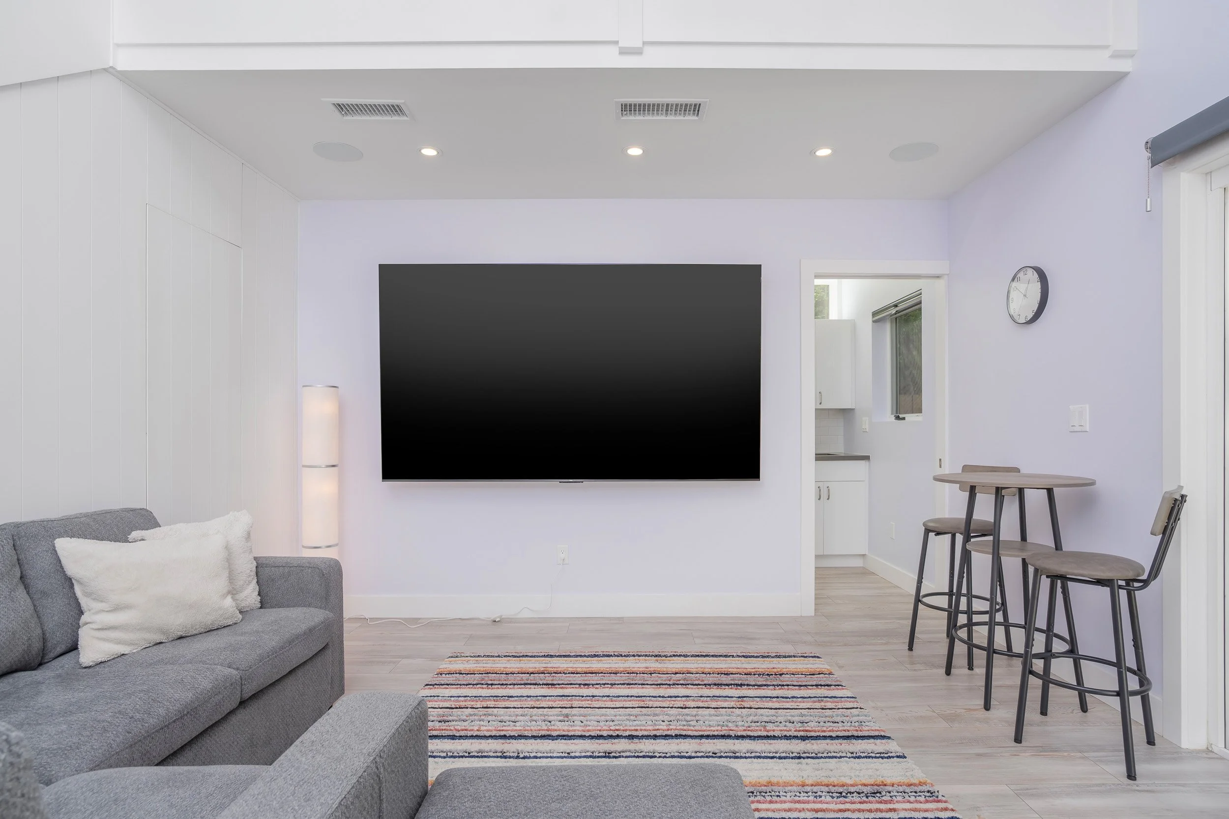 Living room with gray sofa, white pillows, a colorful striped rug, mounted black TV, kitchenette in the background, wall clock, bar stools, and partial view of a kitchen.