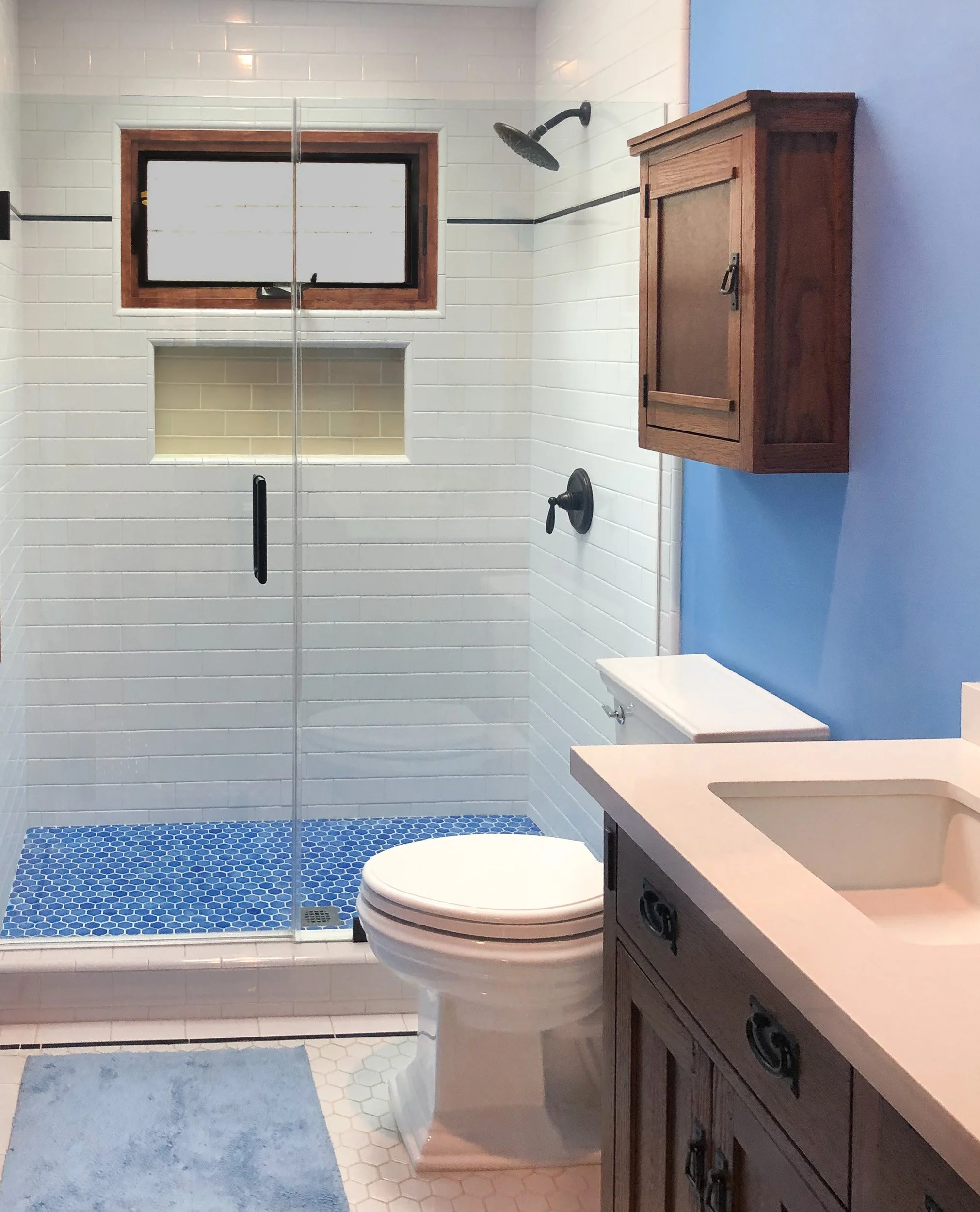 A bathroom with a frosted glass shower door, a wall-mounted showerhead, a white toilet, a wooden cabinet on the wall, a blue accent wall, and a light-colored countertop with a built-in sink. The floor has small hexagonal tiles, and there is a window 
