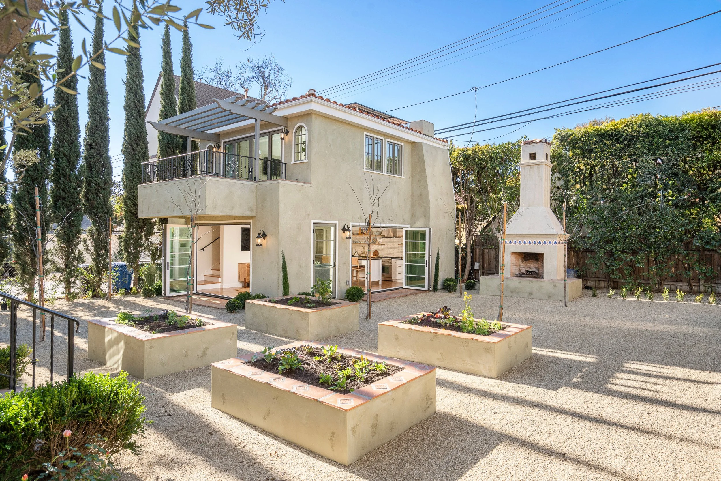 A two-story house with a beige exterior, large windows, and a balcony, next to a tall white outdoor fireplace with a chimney. The yard has raised garden beds with small plants, a gravel pathway, and is enclosed by a wooden fence and lush trees.