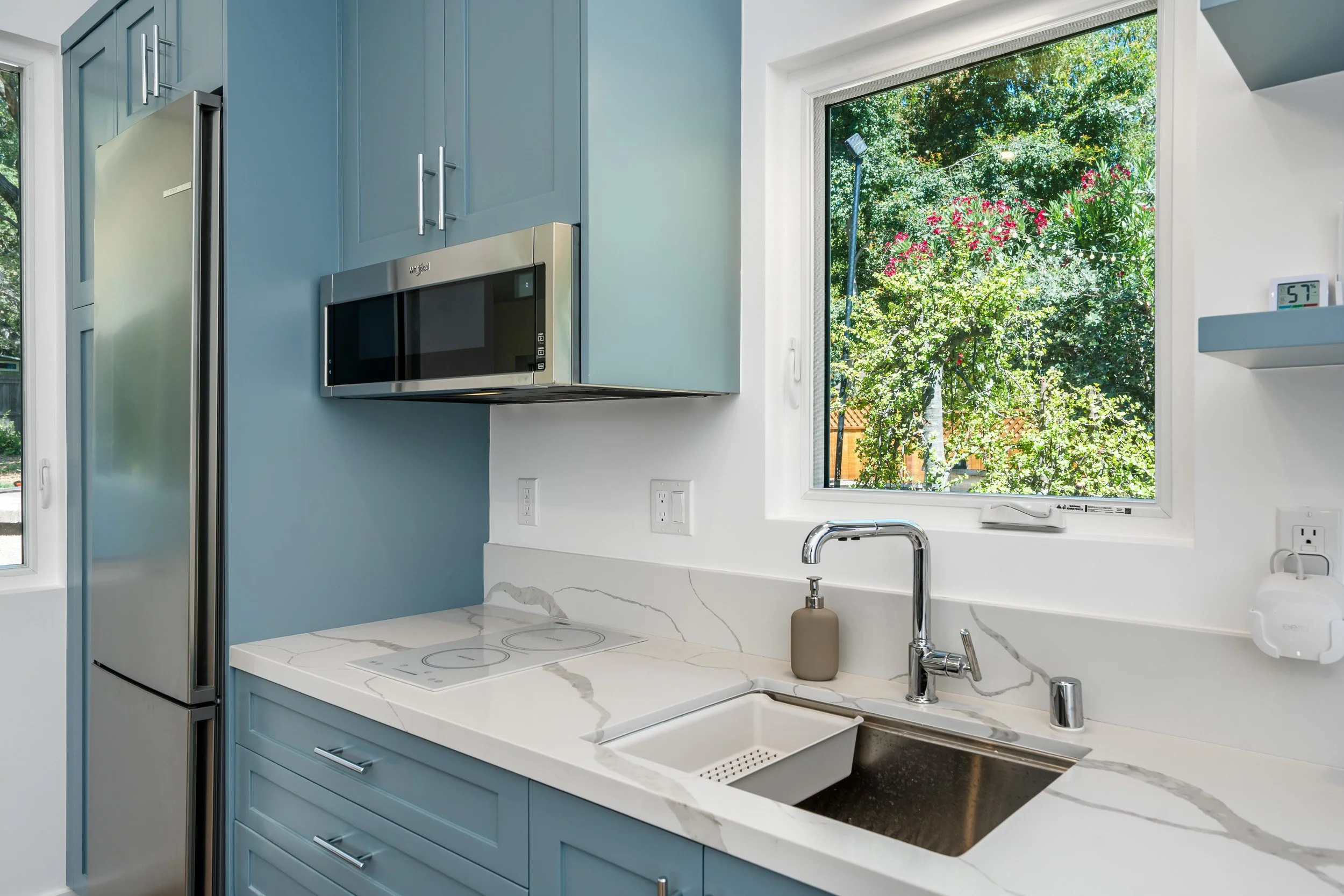 Kitchen with blue cabinets, stainless steel refrigerator, microwave, white marble countertops, window revealing green outdoor foliage, and a white digital thermostat on the wall.