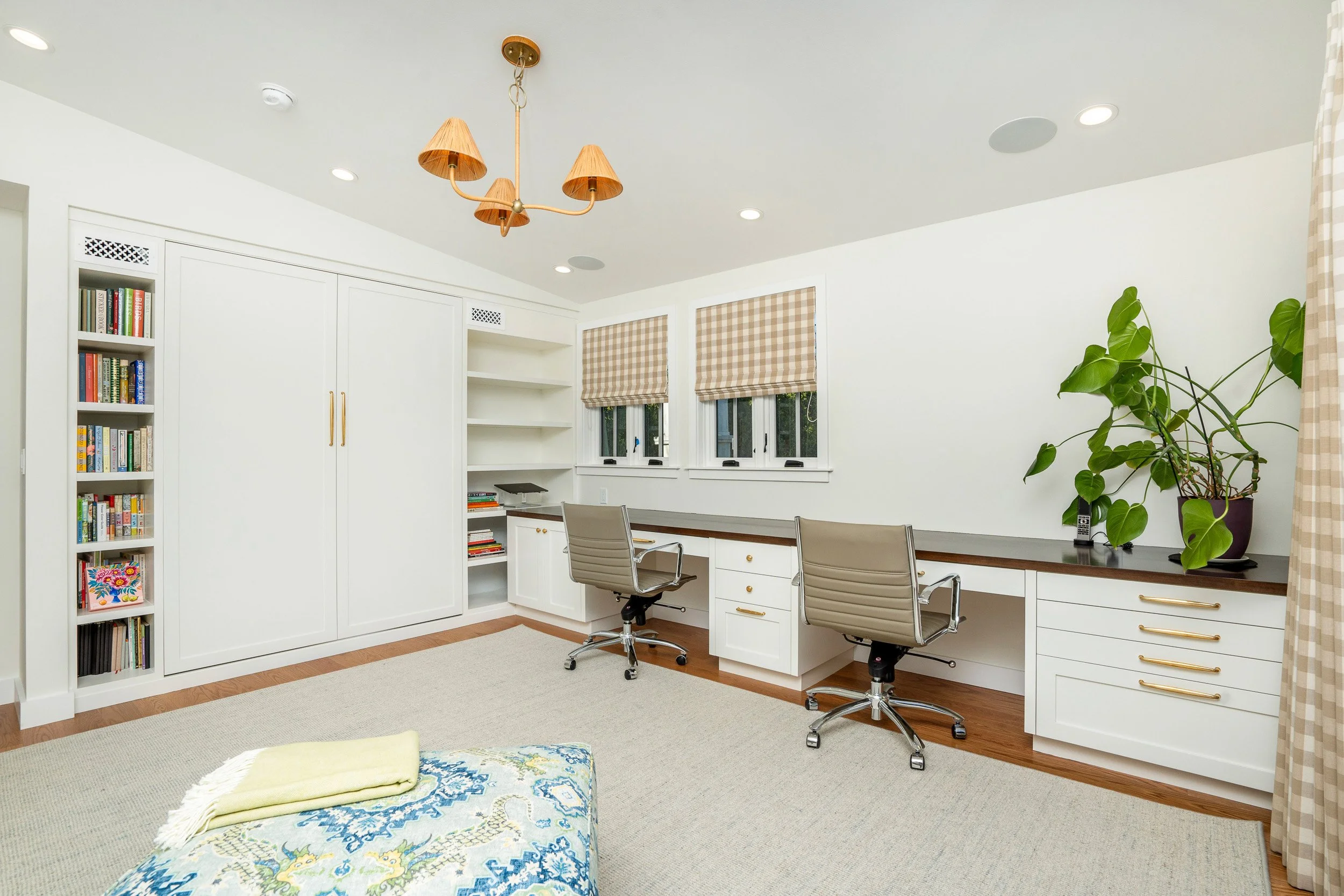 Home office with white built-in shelves, a desk with two beige office chairs, windows with beige checkered blinds, a large green plant, and a ceiling chandelier with multiple shade-like fixtures.
