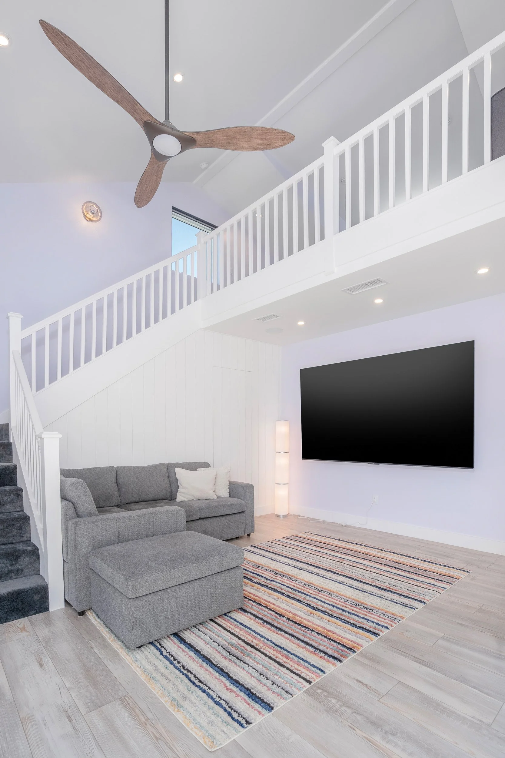 Living room with gray sectional sofa, colorful striped rug, large wall-mounted TV, white walls, staircase with white railing, ceiling fan with wooden blades, and ceiling lights.