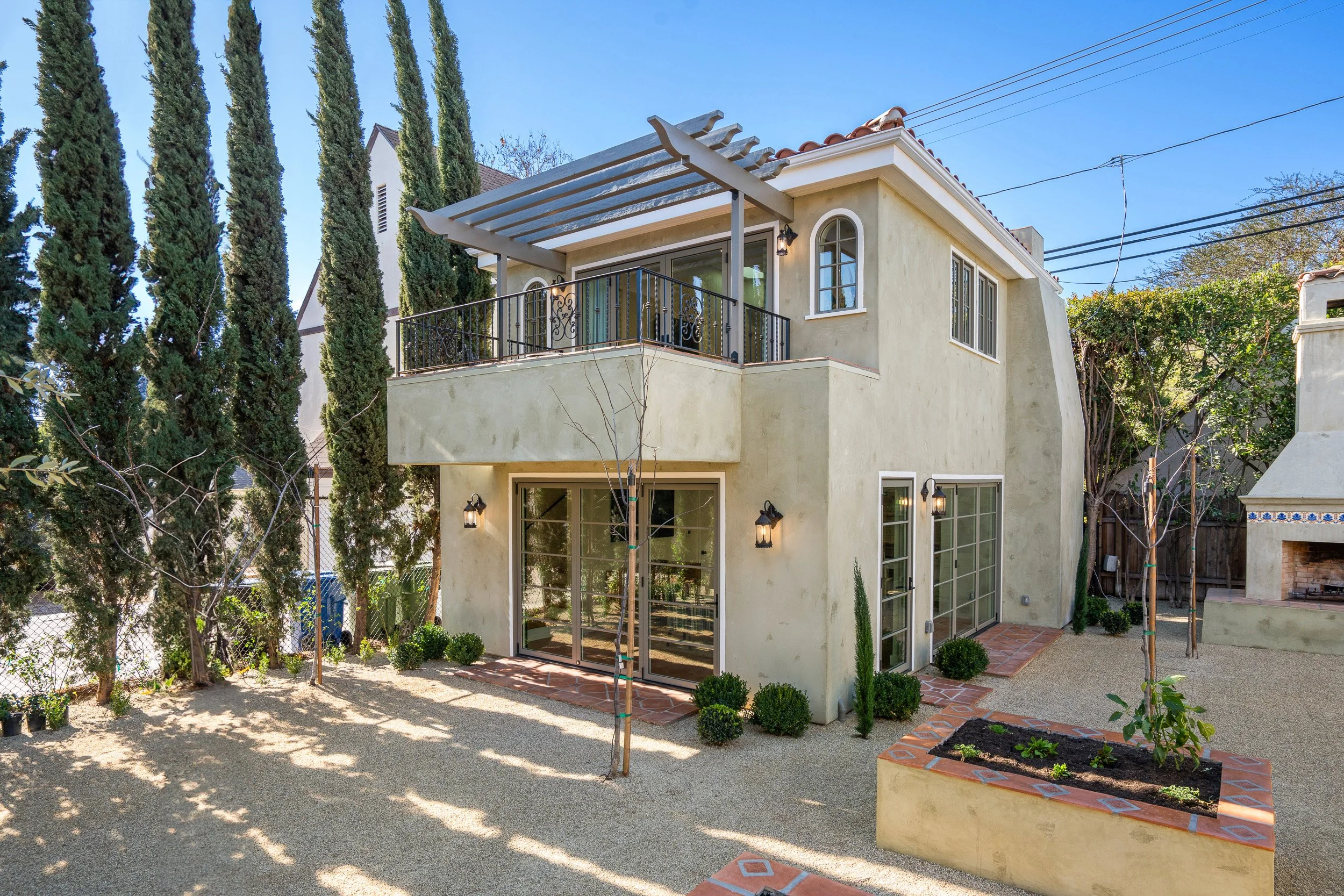 Backyard view of a two-story house with a balcony, large windows, and an outdoor fireplace. Tall cypress trees line the left side, and a small vegetable garden is in the foreground. The sky is clear and blue.