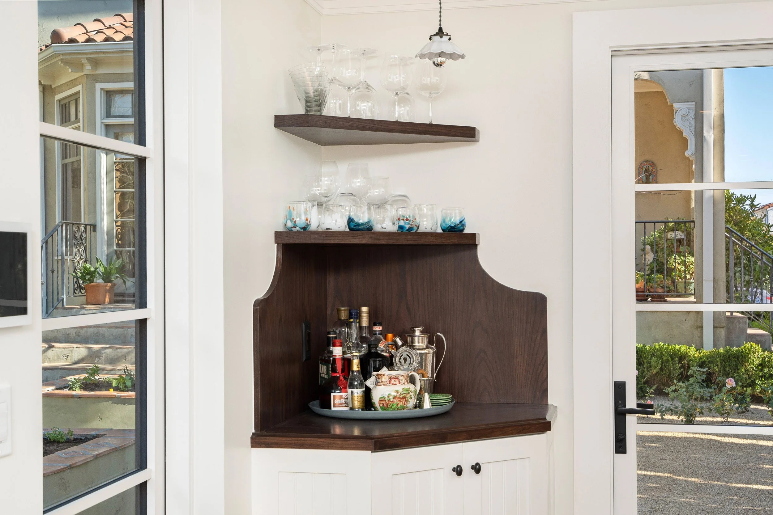 A kitchen interior viewed through a window and glass door. Inside, there is a wooden bar with bottles and barware, and shelves with glassware on the wall.