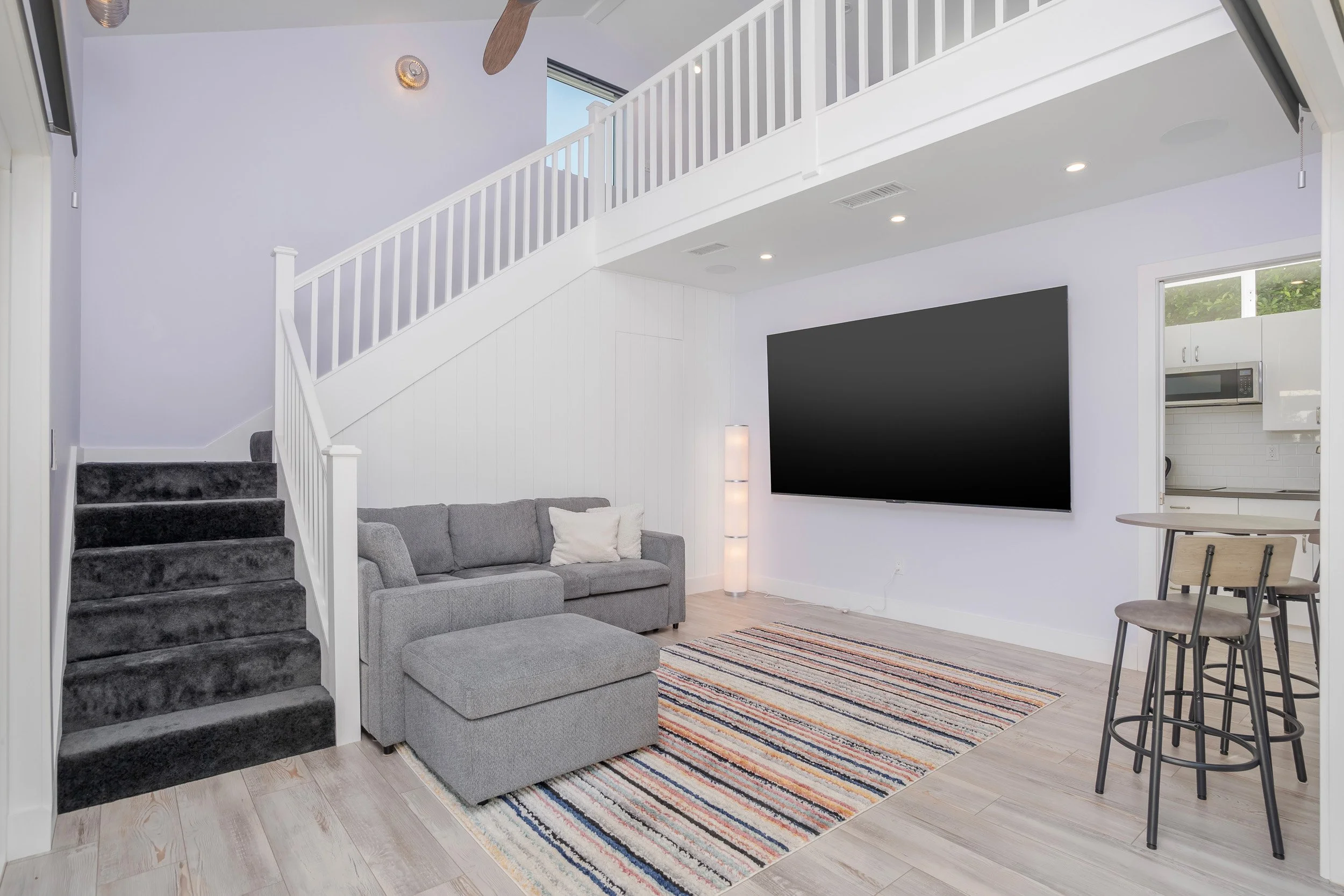 Living room with gray sectional sofa, striped rug, black TV mounted on white wall, staircase with dark carpet, tall floor lamp, and kitchen area visible in background with microwave and cabinets.
