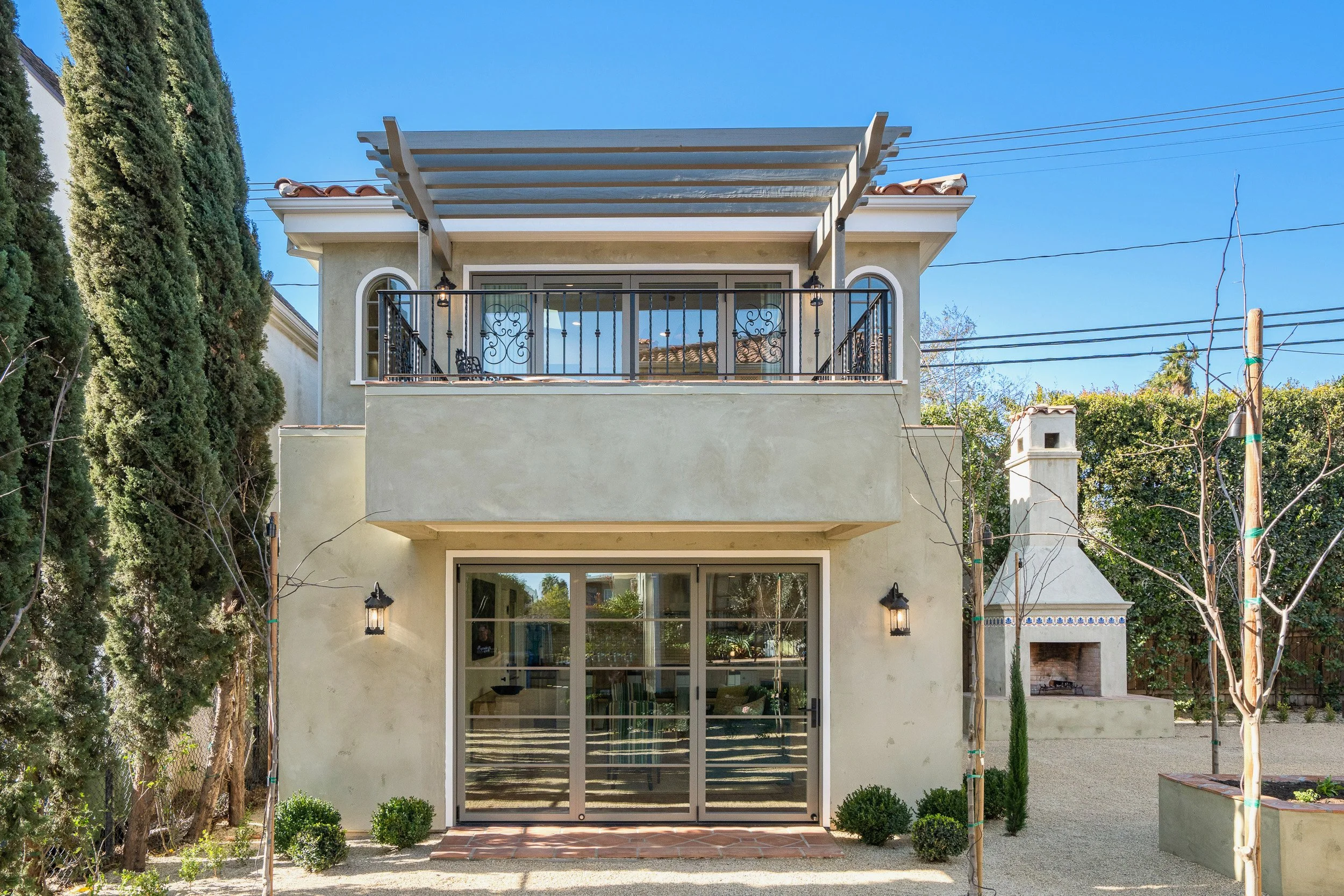 Two-story house with a balcony, sliding glass doors, and outdoor lighting. The balcony has wrought-iron railings and an overhead pergola. Tall trees flank the house, and a firepit is visible on the backyard patio.