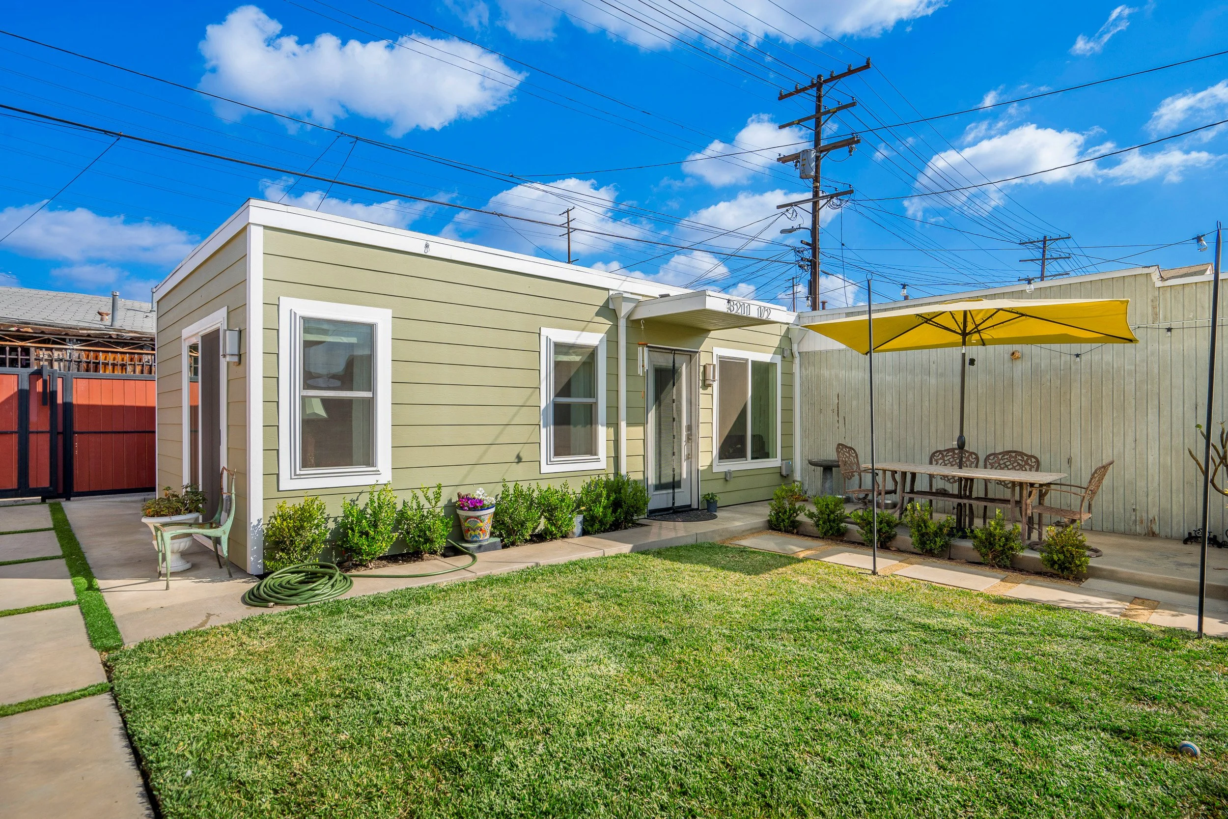 A backyard with a green house, patio furniture, yellow umbrella, and garden along with utility poles and power lines in the background under a blue sky with clouds.