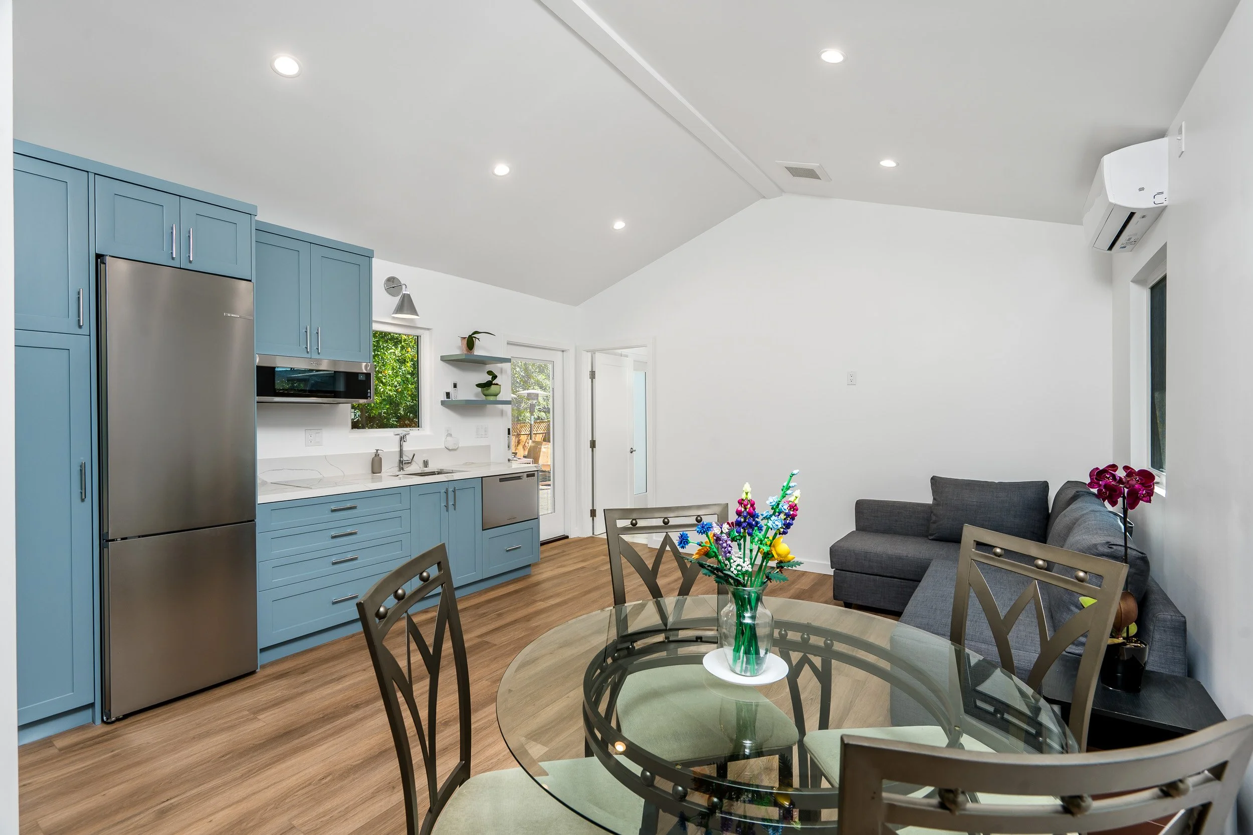 A modern, open-concept living and kitchen area with white walls, wooden floors, and ceiling with recessed lighting. The kitchen has blue cabinets, a stainless steel refrigerator, and a microwave. There's a dining table with a floral centerpiece, and 