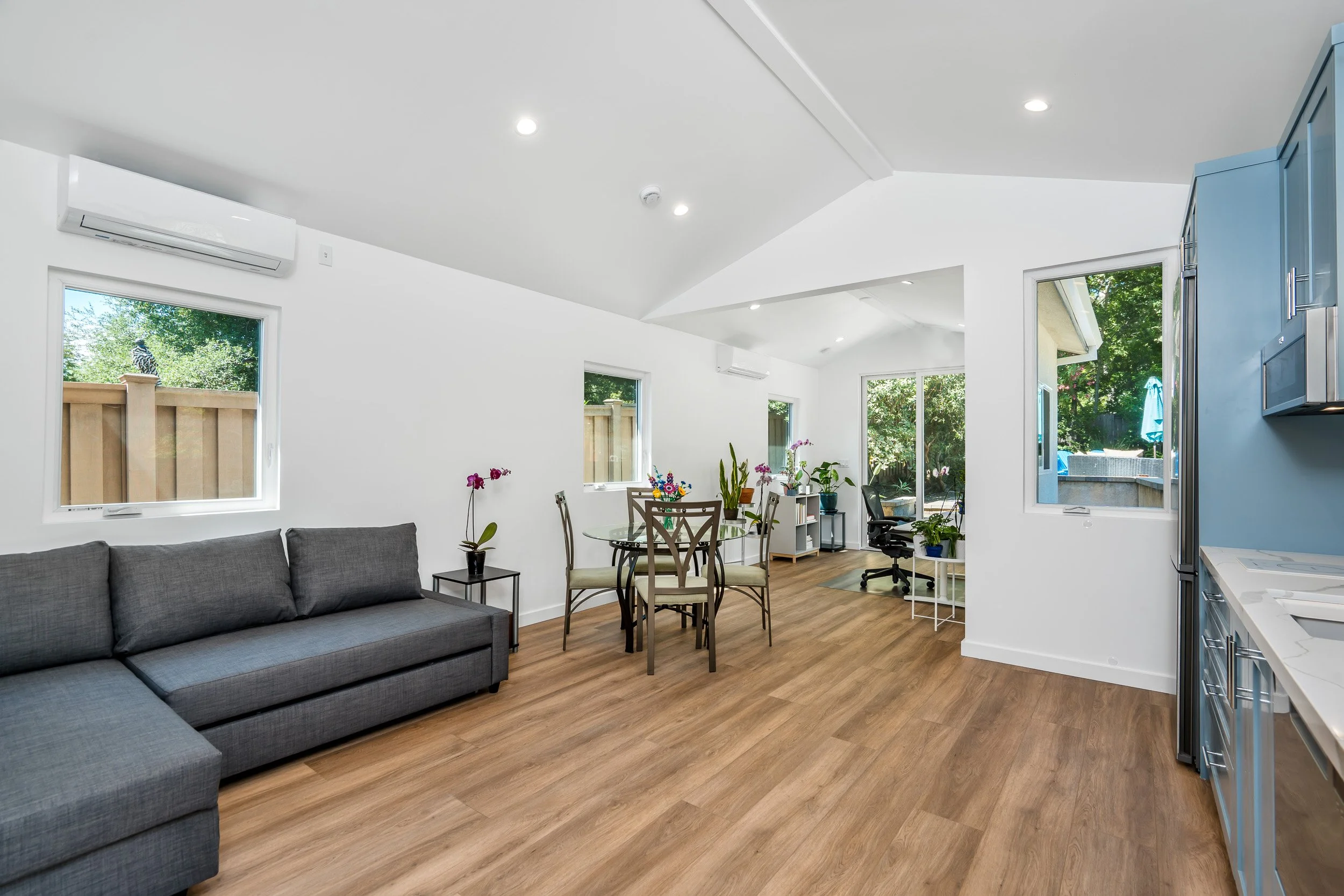 Living room with grey couch, wooden dining table with chairs, plants, large windows showing outdoors, and kitchen with blue cabinets.