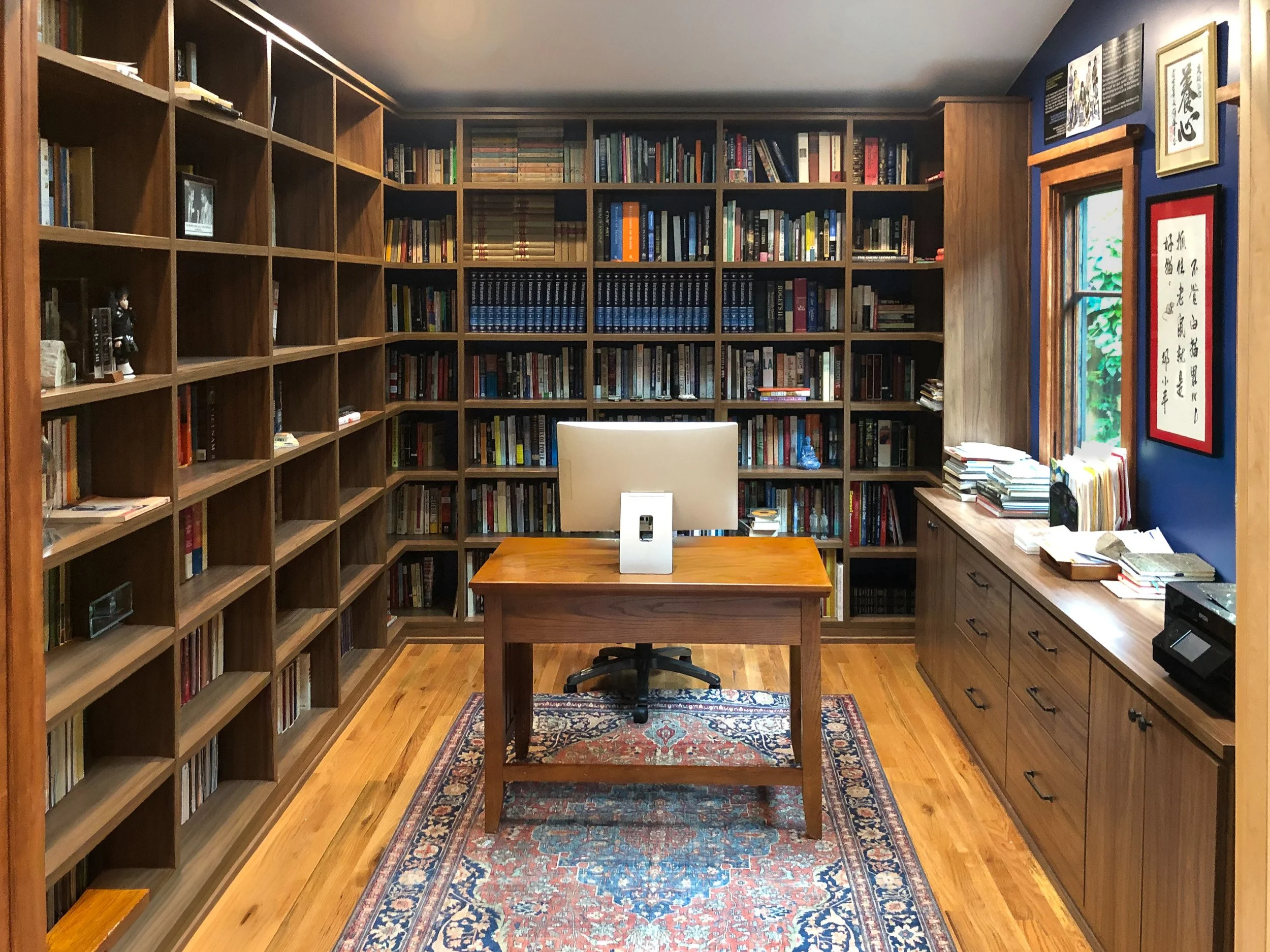 A cozy home library with wooden bookshelves filled with books on all sides, a central desk with a computer monitor, and a decorative rug on the wooden floor.