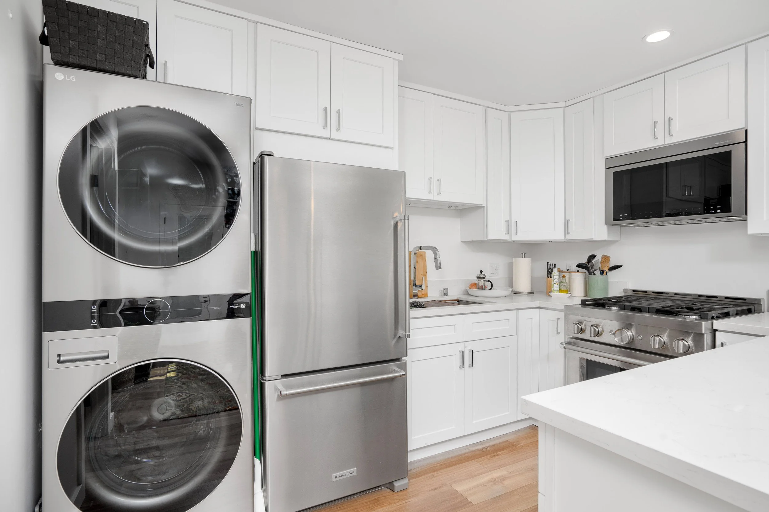 A modern laundry room with white cabinets, a stainless steel refrigerator, a washing machine, and a microwave oven. The countertop has various kitchen items, and the room has hardwood floors.