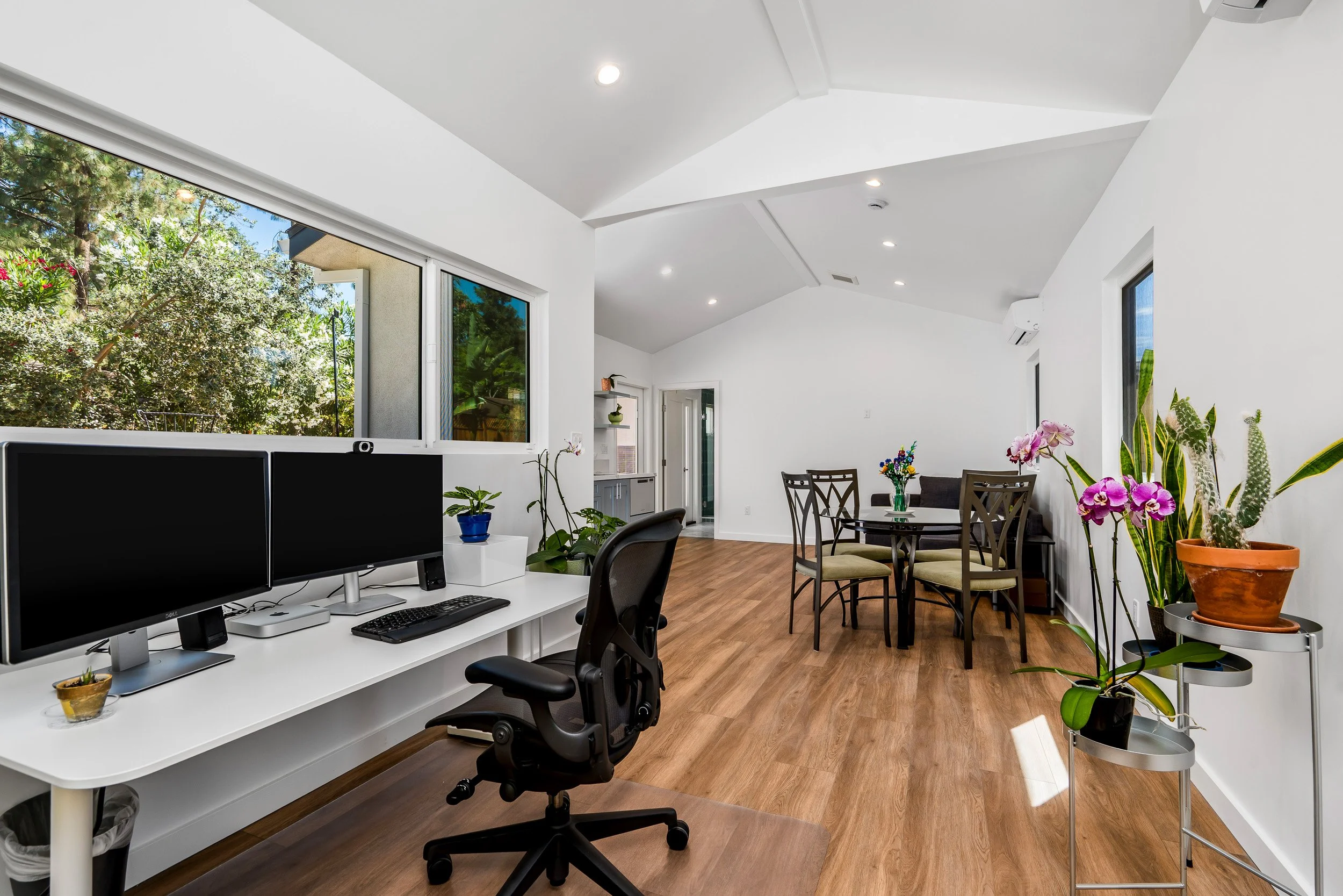 A bright, modern office with a white desk holding two computer monitors, an office chair, and small potted plants. There is a dining table with chairs, several large plants, and windows showing an outdoor view of trees and sky, with recessed ceiling 