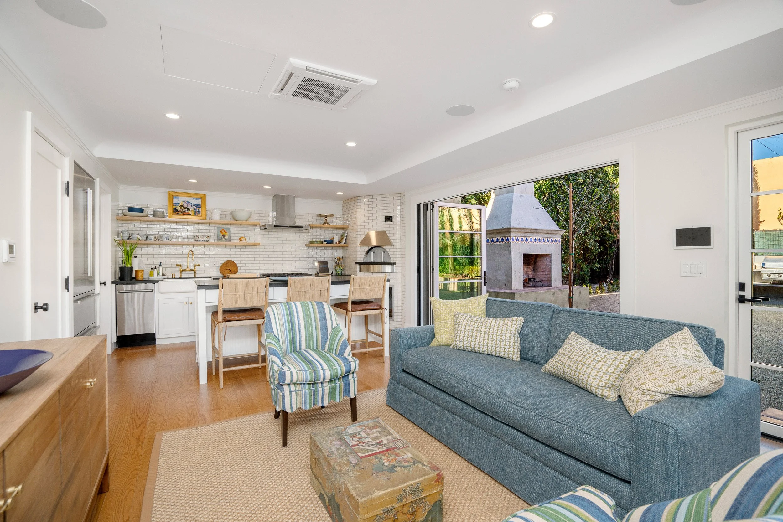Open-concept living room with blue sofa, striped armchair, wooden coffee table, and kitchen with open shelves and outdoor fireplace visible through sliding glass door.