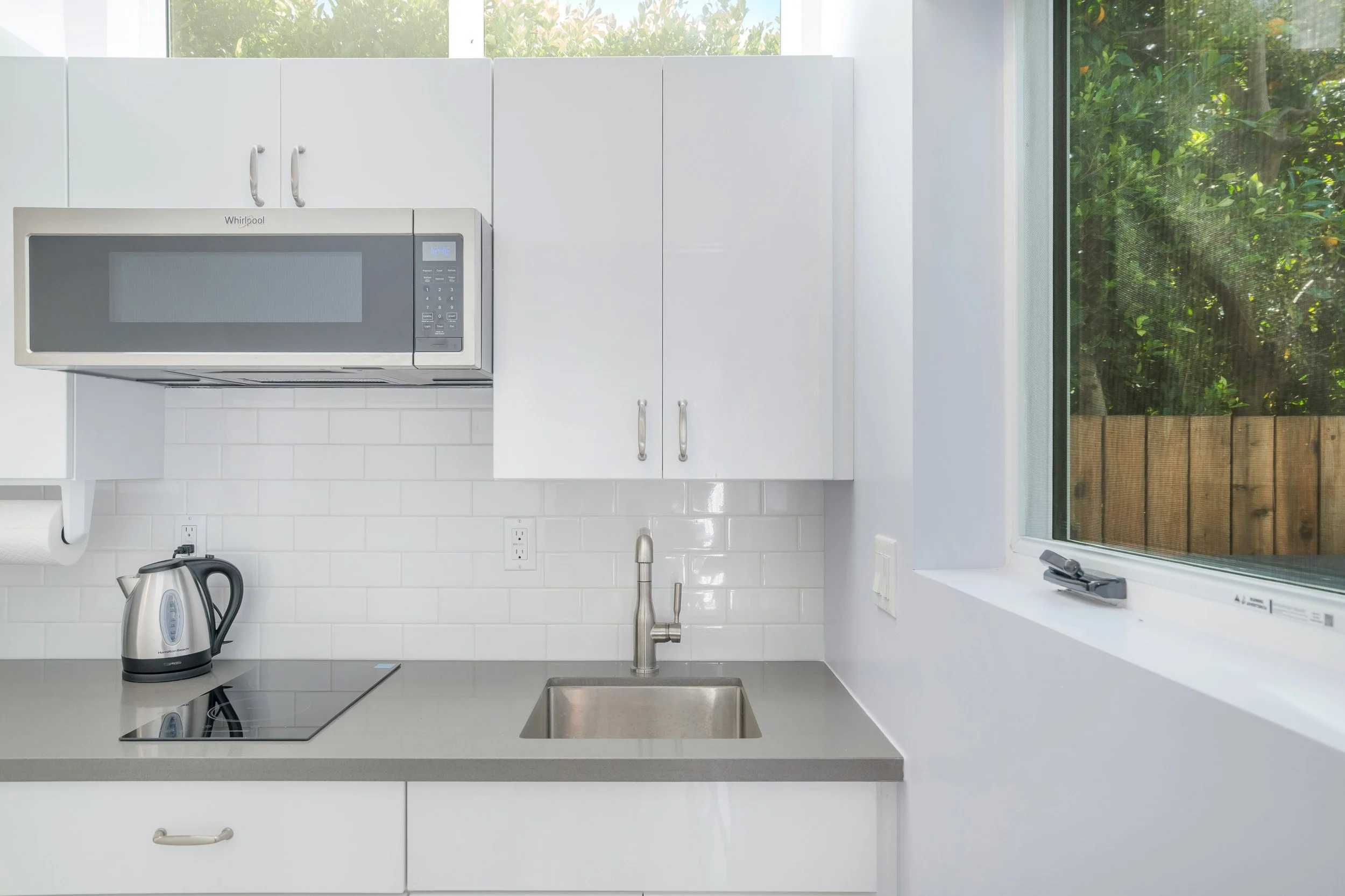 Modern kitchen with white cabinets, a microwave, electric stovetop, electric kettle, sink, and a large window with greenery outside.