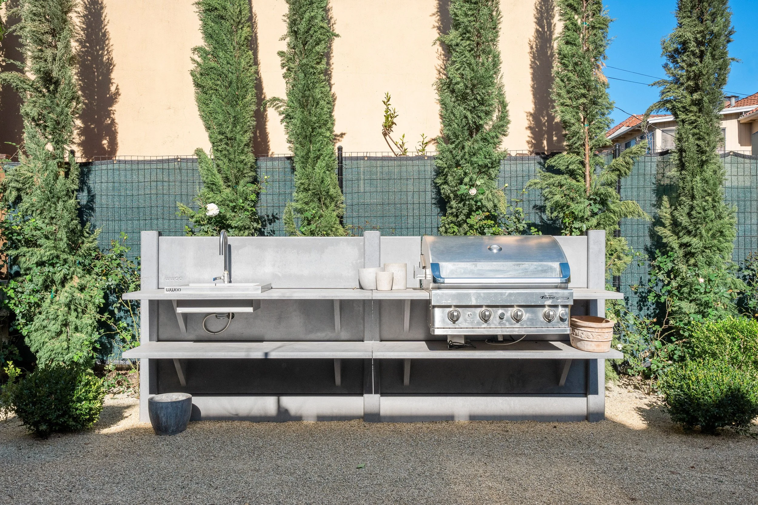 An outdoor kitchen area with a stainless steel grill, a countertop with sink, and potted plants. Tall evergreen trees and a beige wall are in the background.