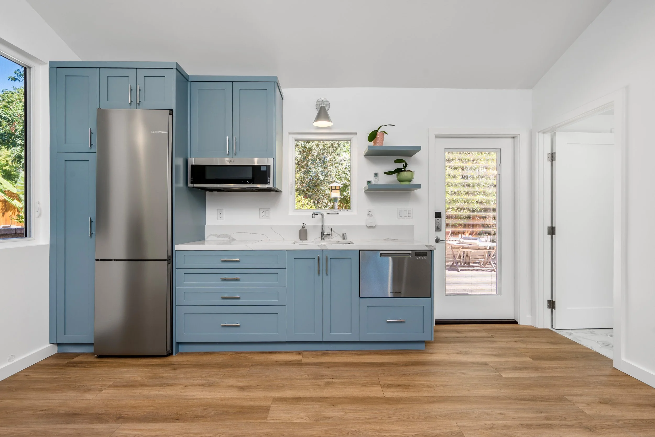 A modern kitchen with blue cabinets, a stainless steel refrigerator, microwave, and dishwasher. There are two windows and a door leading outside, with a small balcony or deck visible through the door. The kitchen has wood flooring and white walls.
