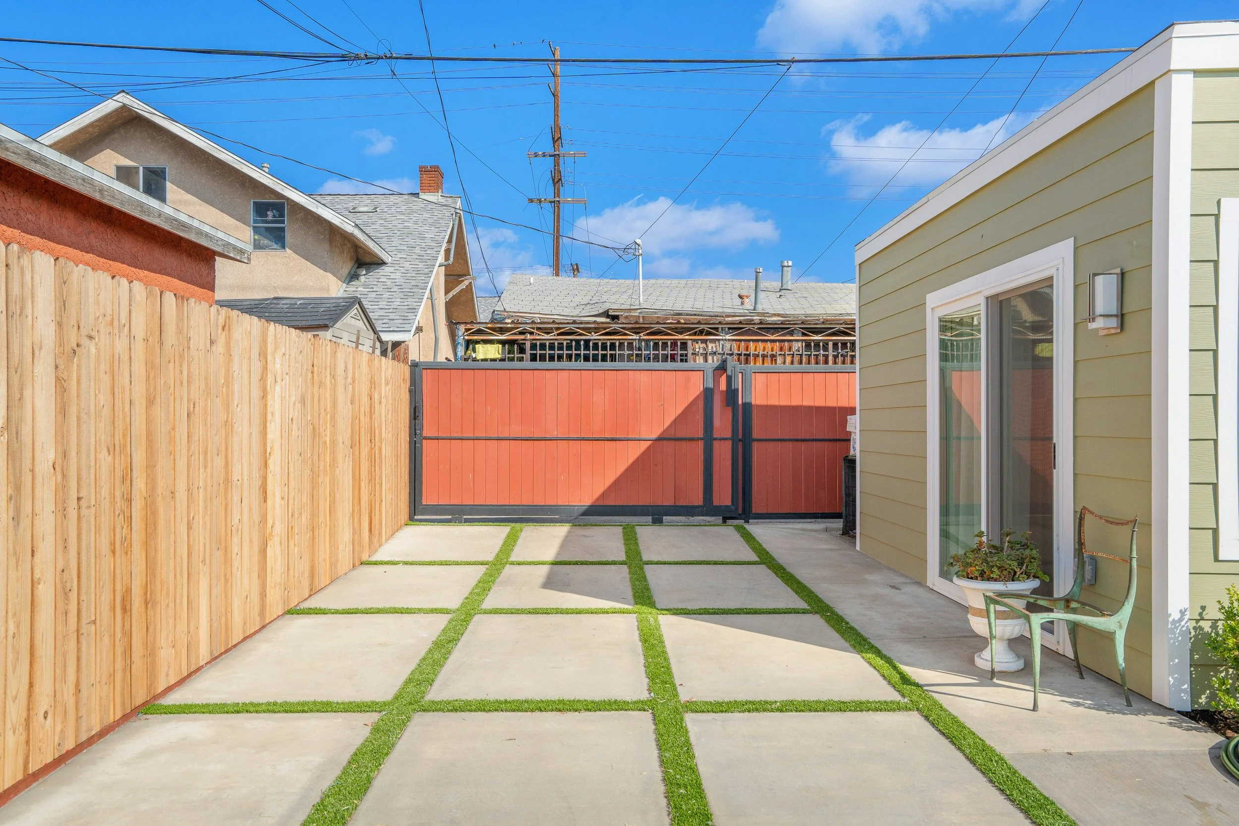 Backyard with concrete patio stones separated by patches of grass, a brown wooden fence on the left, and a green house with a sliding glass door on the right. A metal chair and a potted plant are next to the house. A red gate is in the middle, and ne