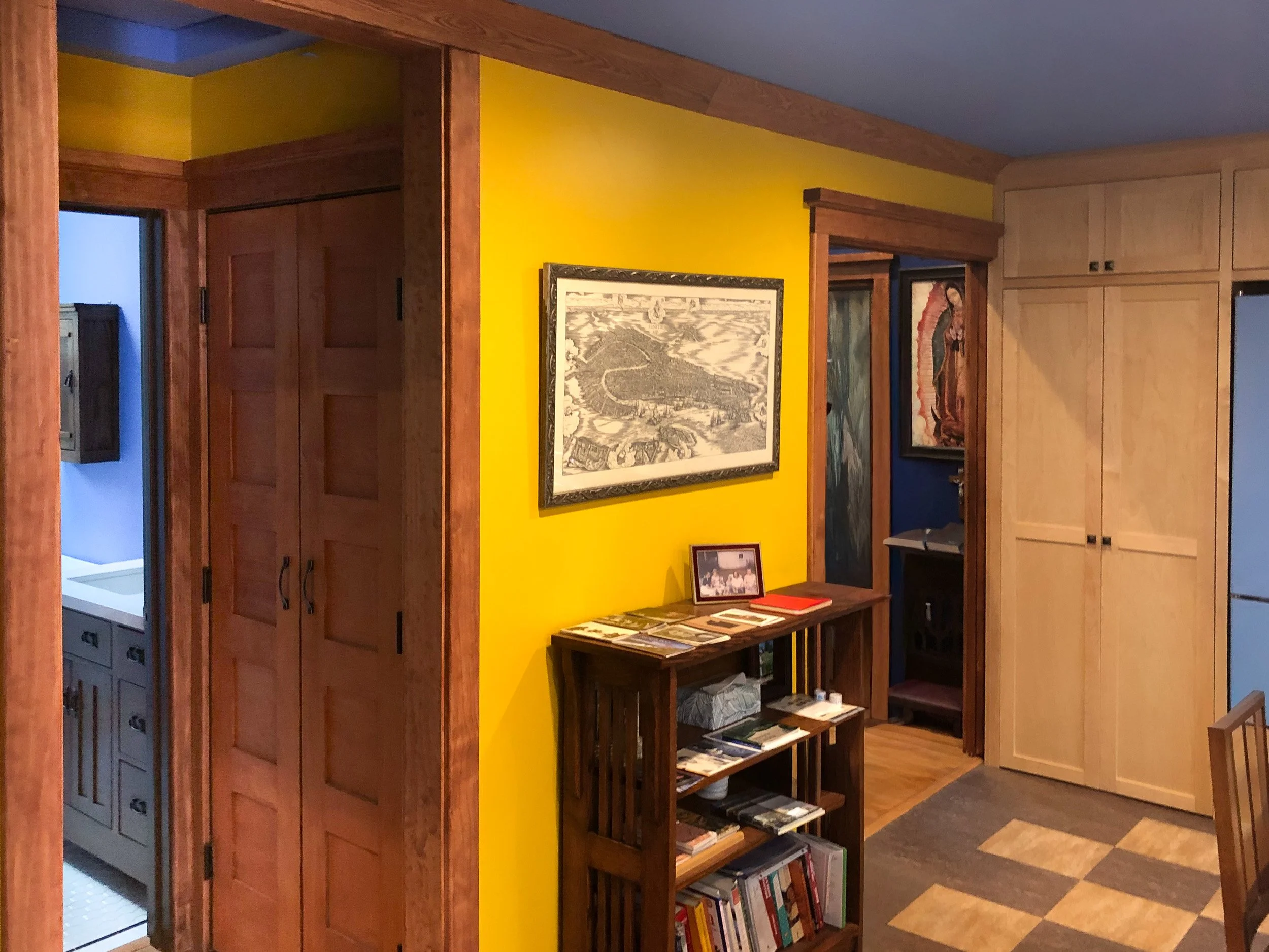 A cozy interior corner with wooden shelves filled with books, a yellow wall, and framed artwork. There is a doorway leading to another room with blue walls.