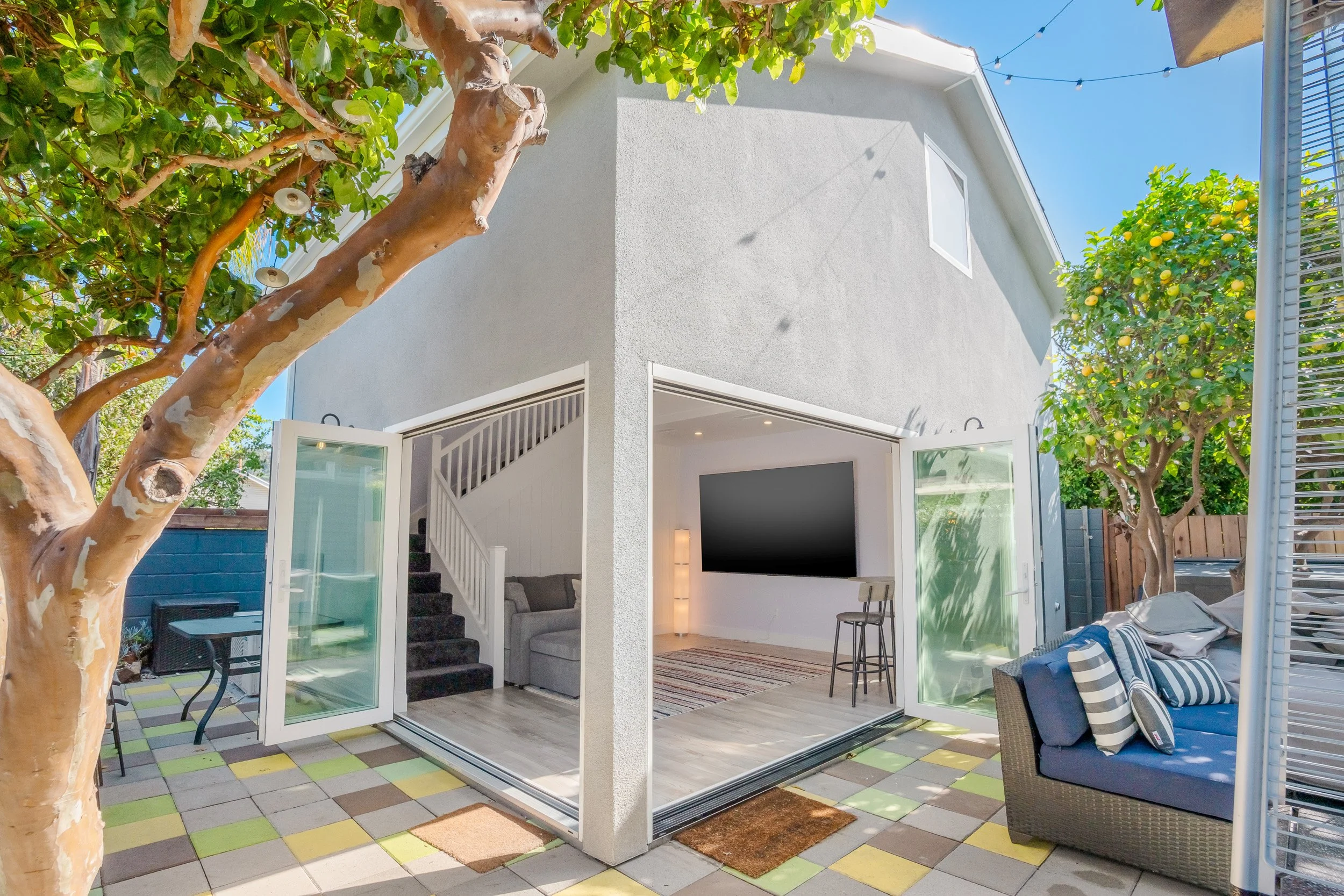 A backyard patio with an open sliding glass door leading into a living room. There are trees with lemon-like fruit and string lights overhead. There is outdoor furniture, and inside, a living room with a sofa and a wall-mounted TV. The patio floor ha
