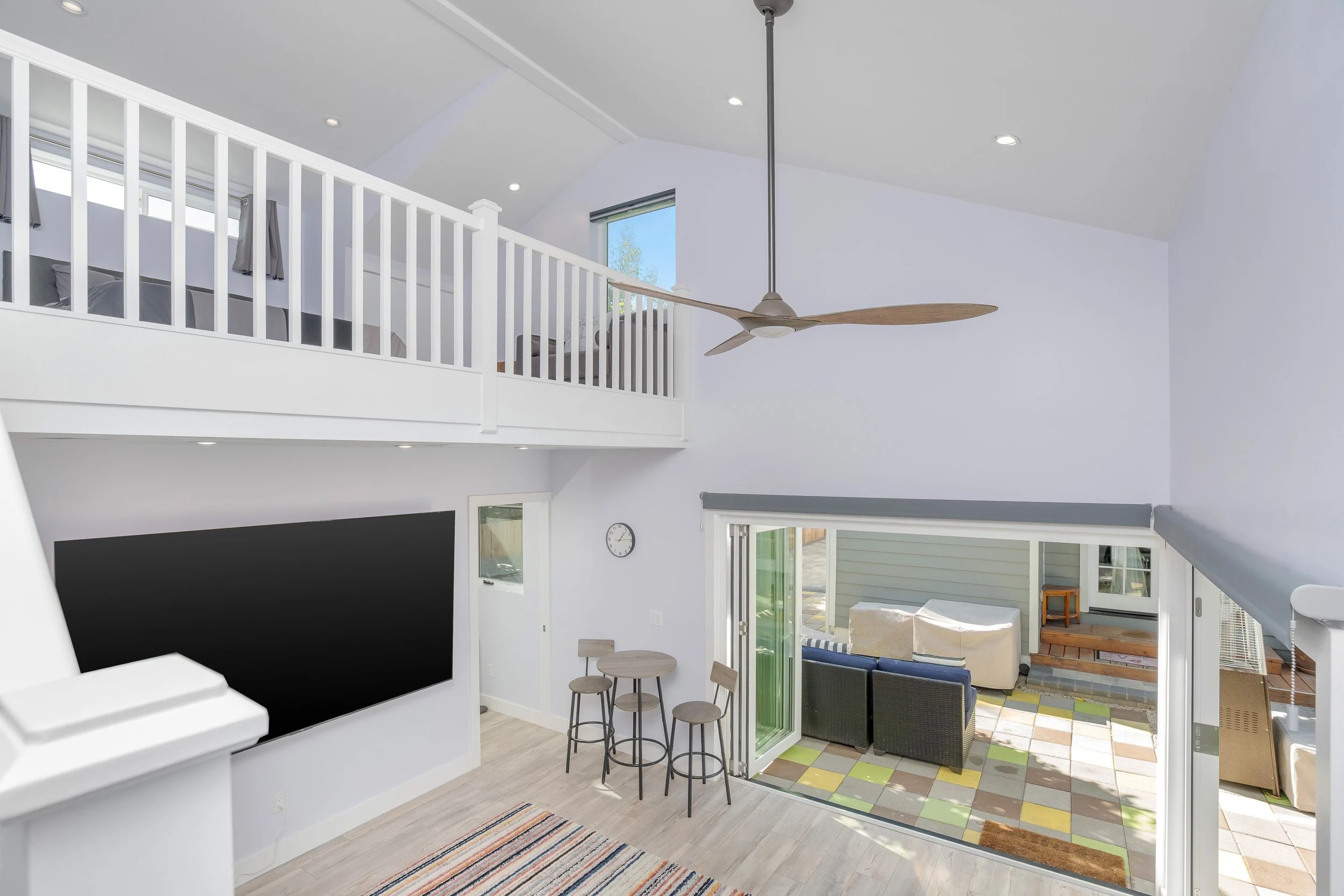 Interior view of a modern, bright living space with white walls, a ceiling fan with wooden blades, a large black chalkboard or TV, a small dining area with three stools, and an outdoor patio area with a sofa and a wooden bench, all with great natural