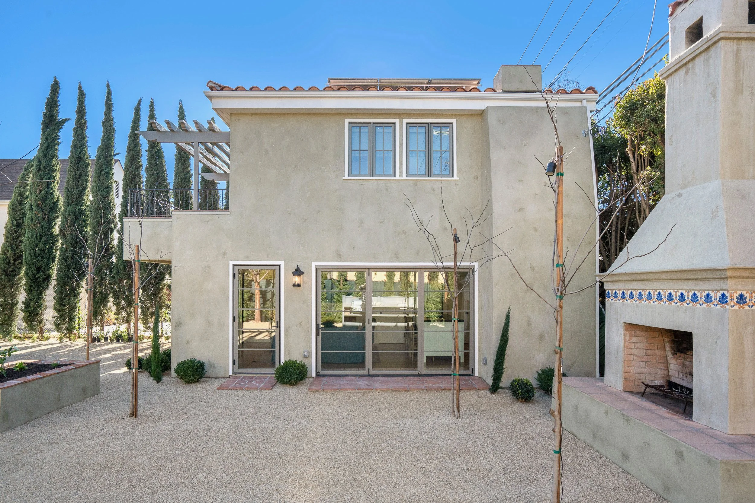 A modern two-story house with large windows, a stucco exterior, and a tiled roof. The backyard has a gravel surface with small shrubs and an outdoor fireplace on the right. Tall cypress trees and utility poles are visible in the background under a cl