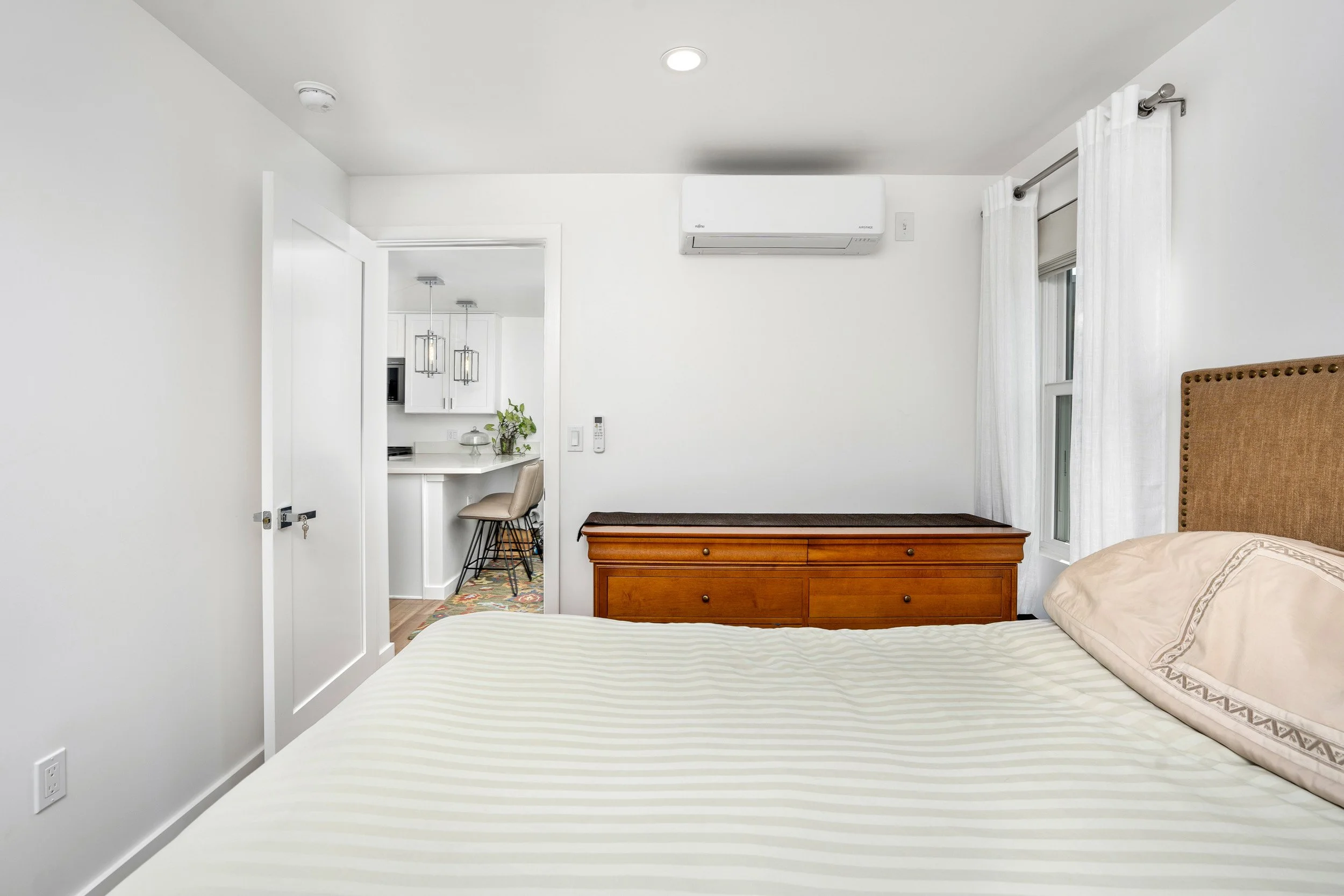 A bedroom with a large bed that has striped bedding, a wooden dresser, a window with white curtains, and a corner of a headboard. There is an opening to a small space with a white kitchen and two bar stools, accented with pendant lights and some deco