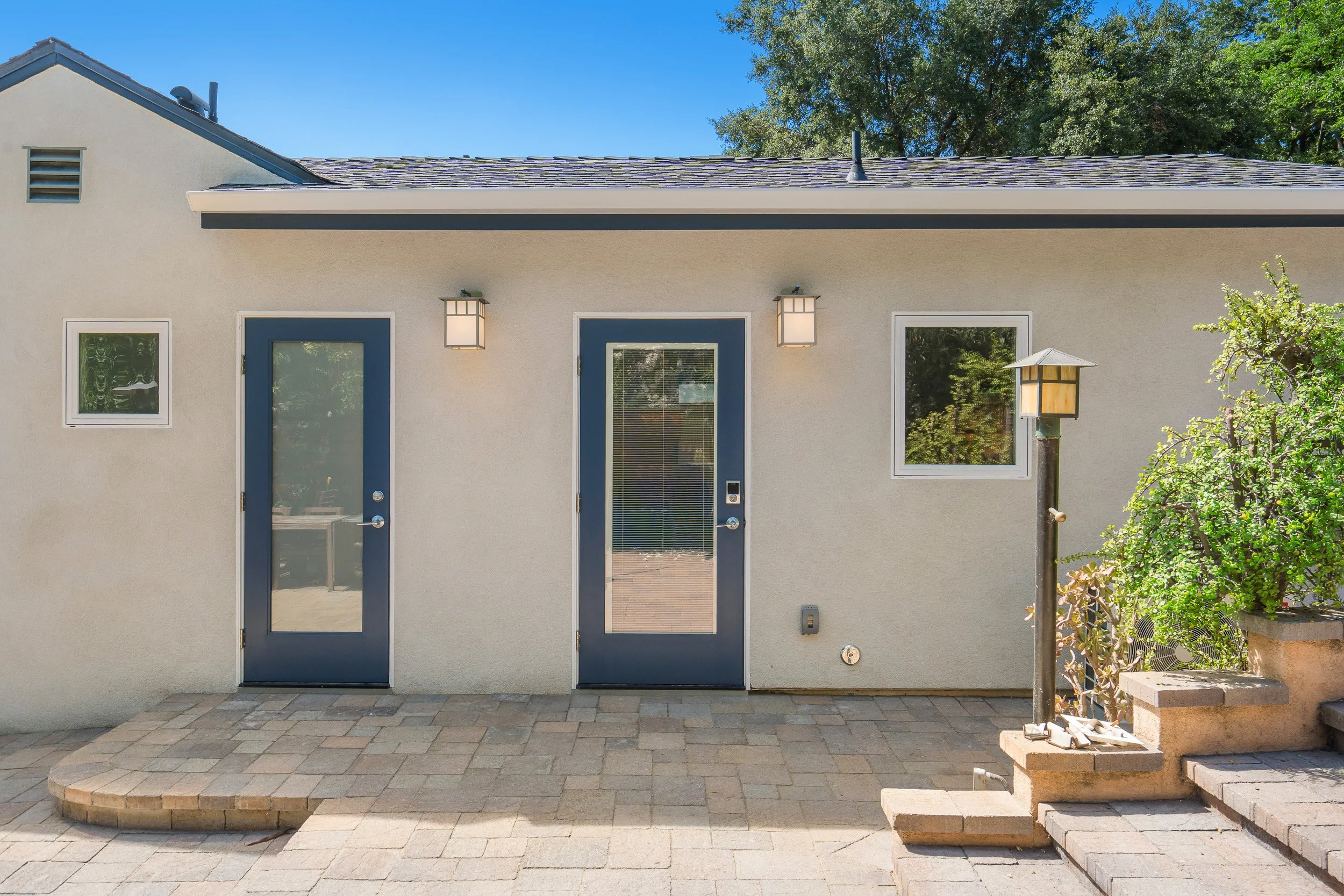 Backyard patio with two blue front doors, two outdoor wall lanterns, windows, brick steps, stone paver patio, and a small tree on the right.