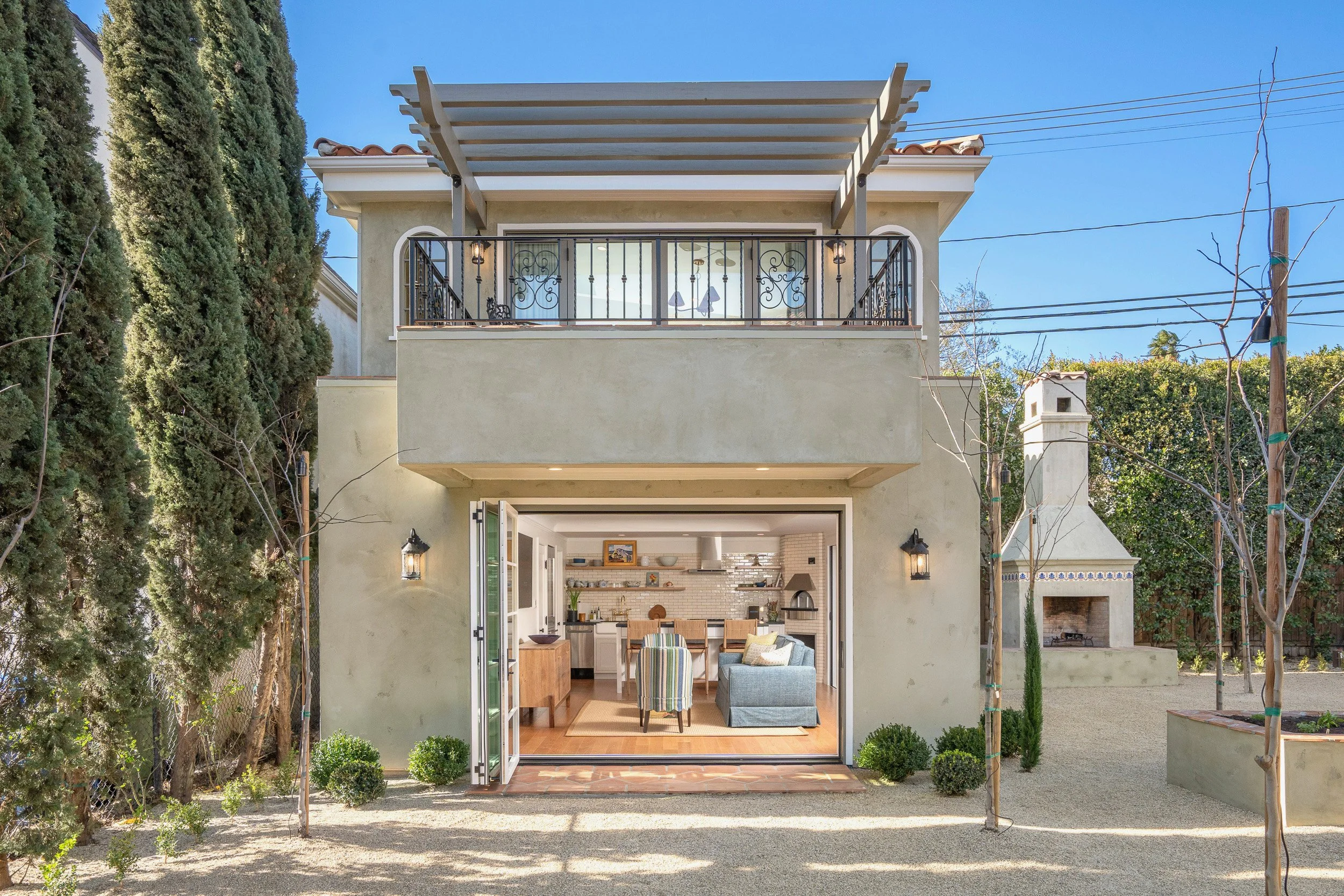 A modern, two-story house with a large open sliding door on the ground level, revealing a cozy, well-lit living room with a white brick fireplace and furniture inside. The second floor features a balcony with wrought-iron railings, lighting fixtures,