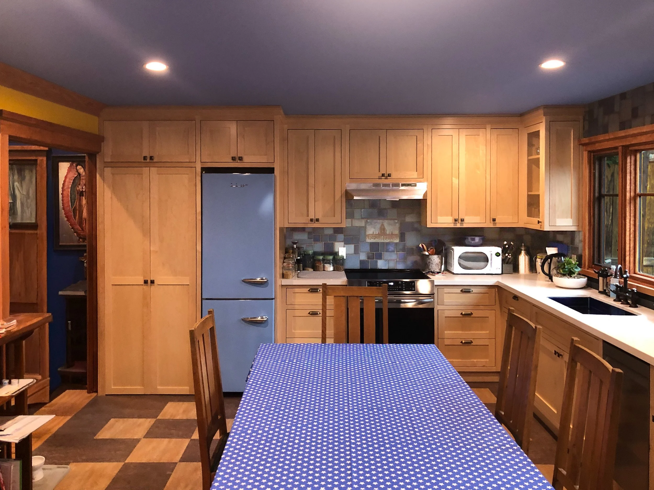 A kitchen with wooden cabinets, a blue refrigerator, a stove, a microwave, and a dining table with a blue tablecloth with white stars. There are windows on the right side and a picture of a fish and a seahorse on the left wall.