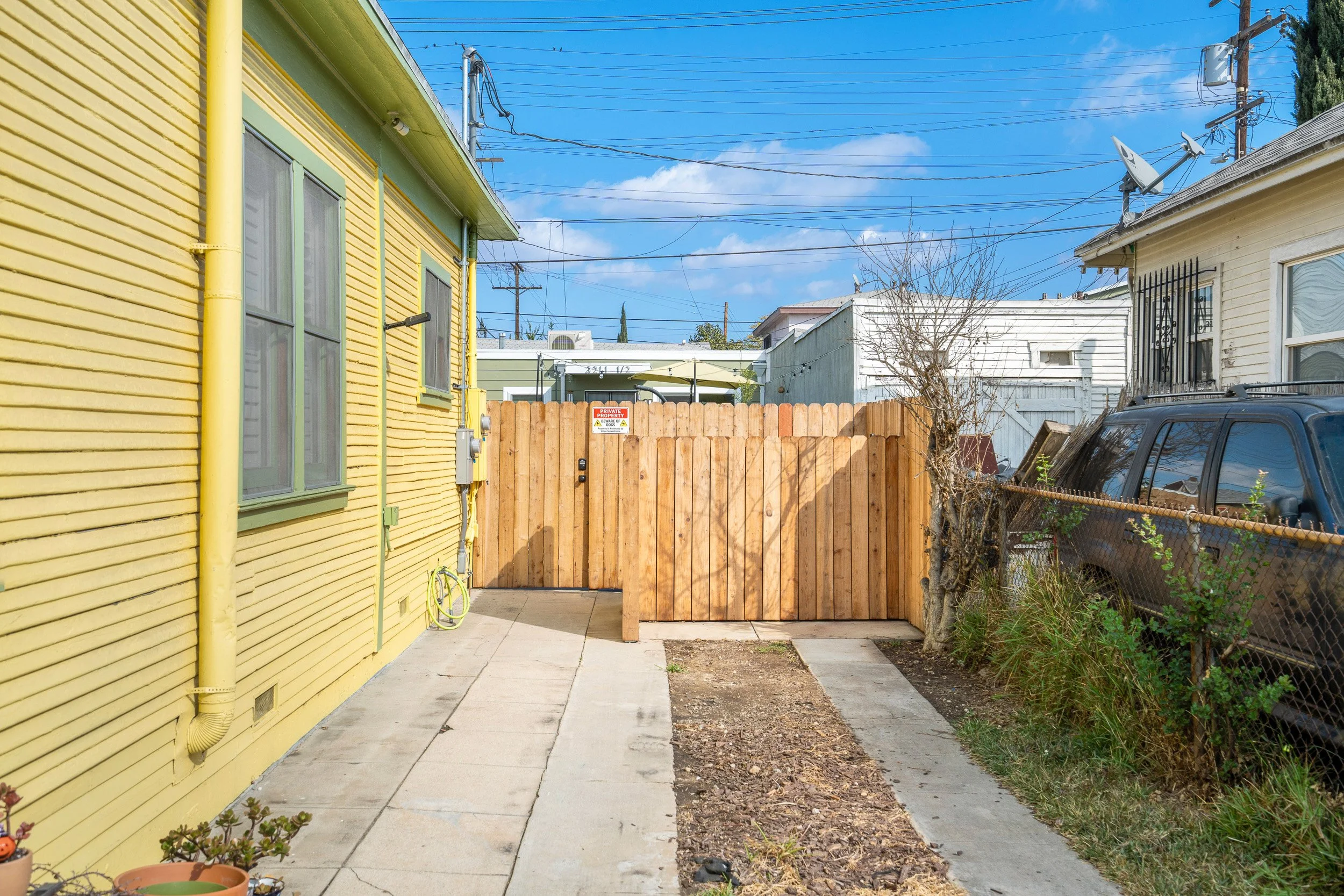 A backyard with a yellow house on the left, a wooden fence in the center, and a white house on the right. The sky is blue with some clouds. There are electric wires and poles overhead, some satellite dishes on the roofs, a parked vehicle, and bare tr