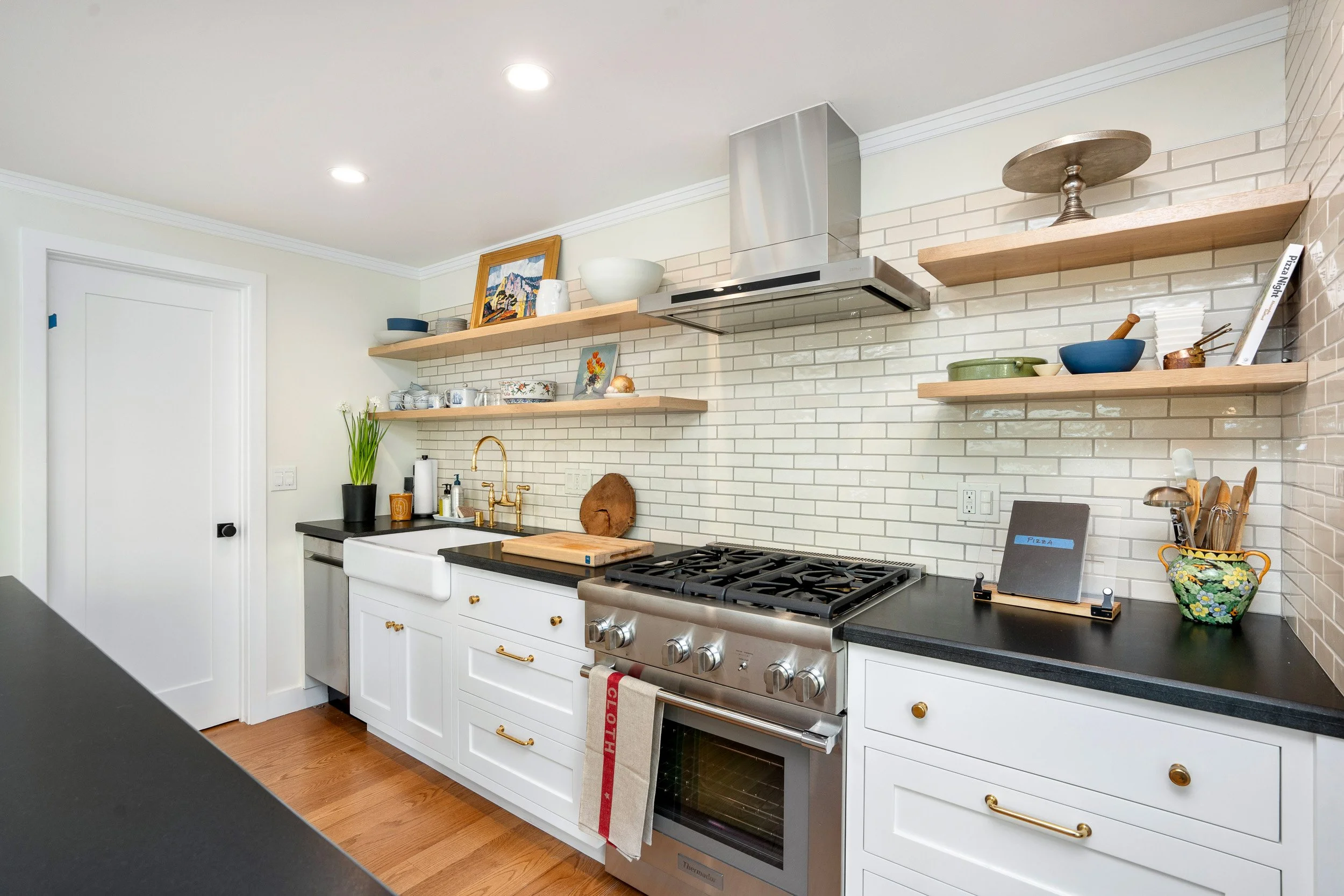 A modern kitchen with white cabinets, floating wooden shelves, a gas stove, a farmhouse sink, and various decorative items and utensils on the counter.