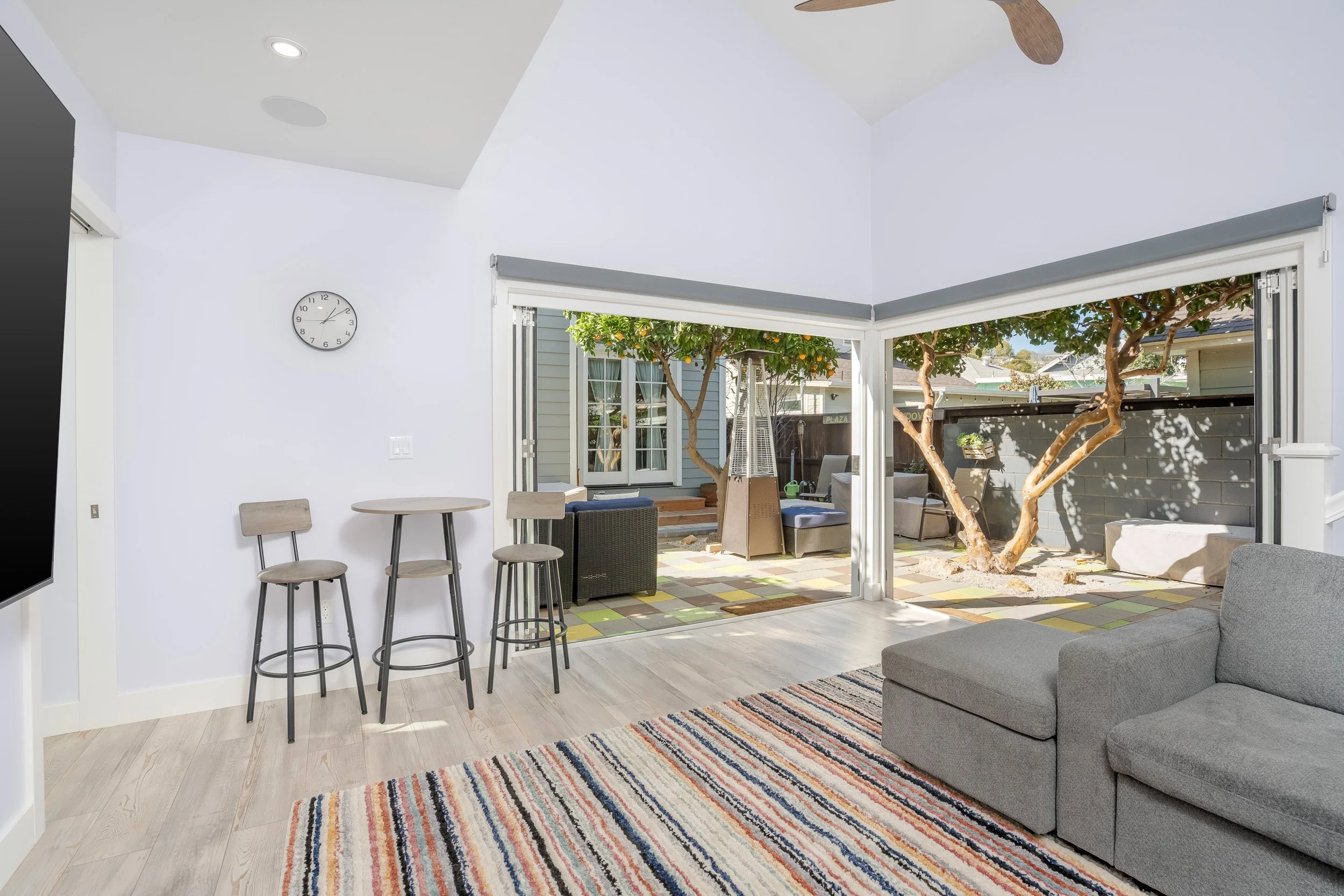 Living room with sliding glass door opening to patio with tree and outdoor furniture, white walls, light hardwood floors, colorful striped rug, gray sofa, round dining table with chairs, wall clock, ceiling fan, and recessed lighting.
