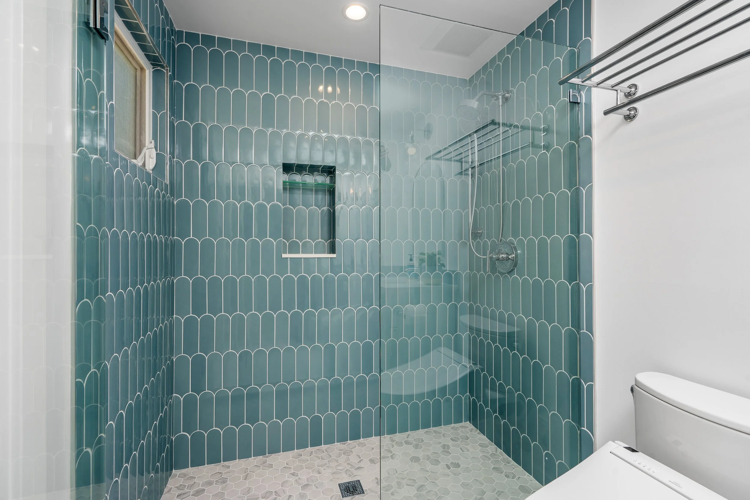 Modern bathroom with teal tiled walls, glass shower enclosure, and white fixtures, including a toilet, with a small window and a wall-mounted towel rack.