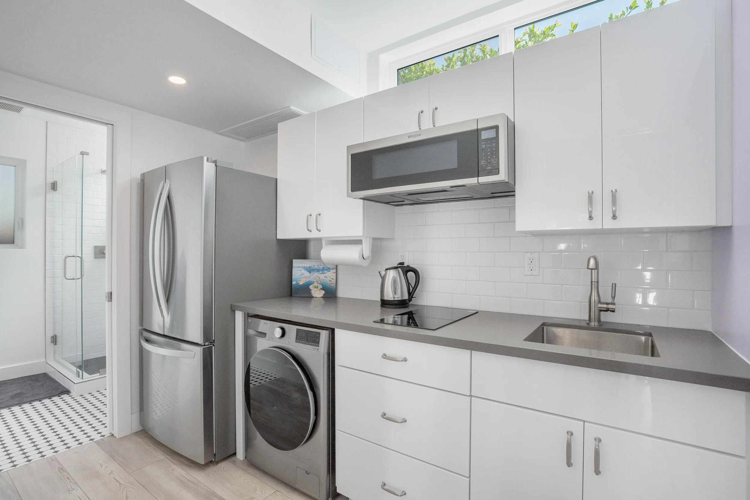 Modern kitchen with white cabinets, stainless steel appliances including a refrigerator, microwave, and washing machine, a black electric stove, and a sink with a window above. There is also a small section of a tiled bathroom or closet door visible 