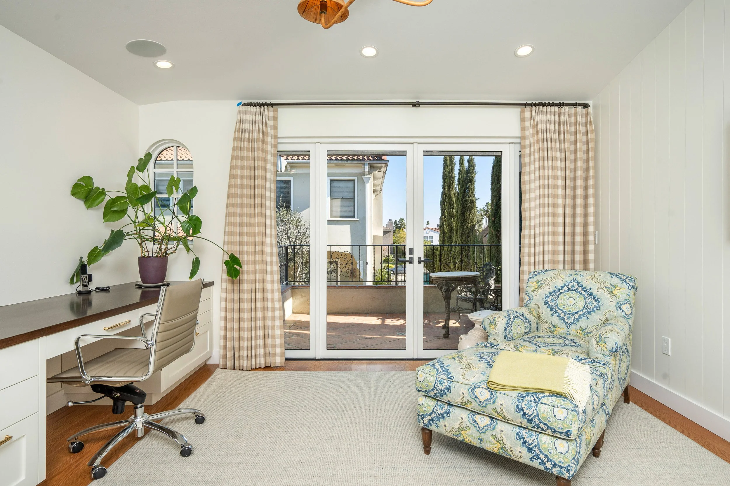 A living room with a sliding glass door leading to a balcony, beige checkered curtains, a white desk with a chair and a potted plant, and a patterned armchair with a yellow blanket. Outside, there are trees and neighboring houses.